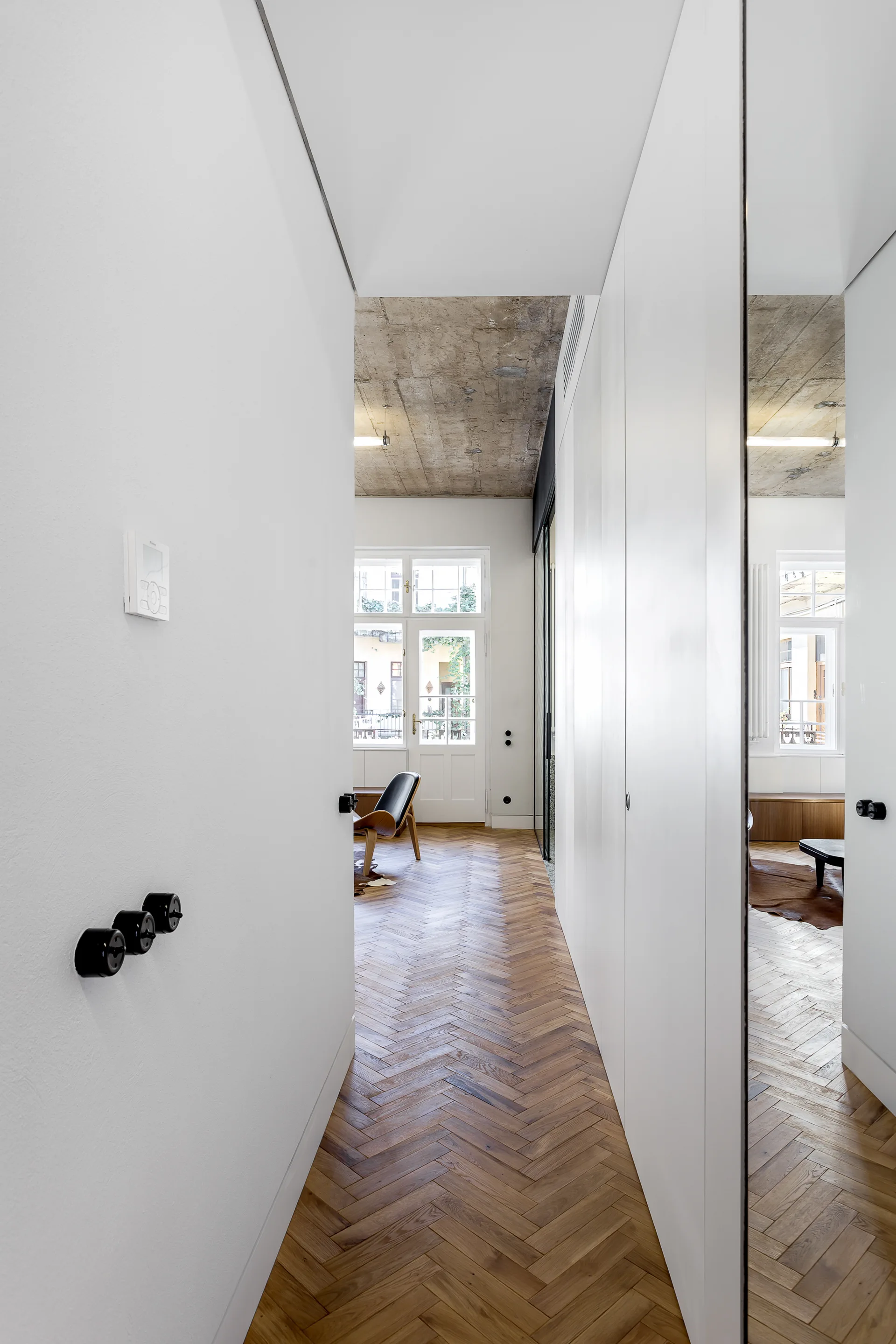 A bright hallway features herringbone hardwood flooring, seamless white wall panels, and a concrete ceiling leading into a light-filled living area.