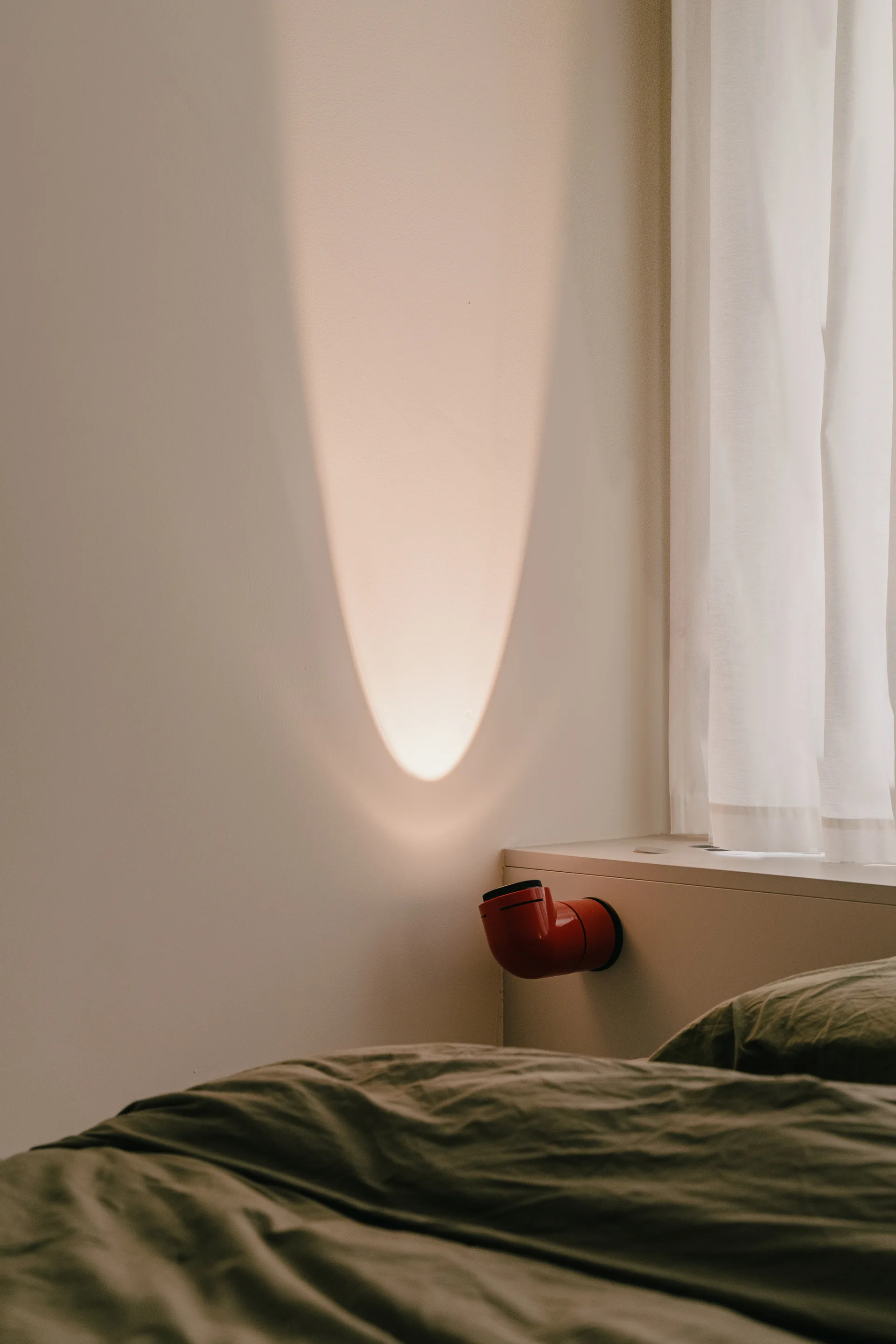 A bright red cylindrical wall lamp is mounted on a beige bedside ledge, casting a warm beam of light onto an off-white wall.