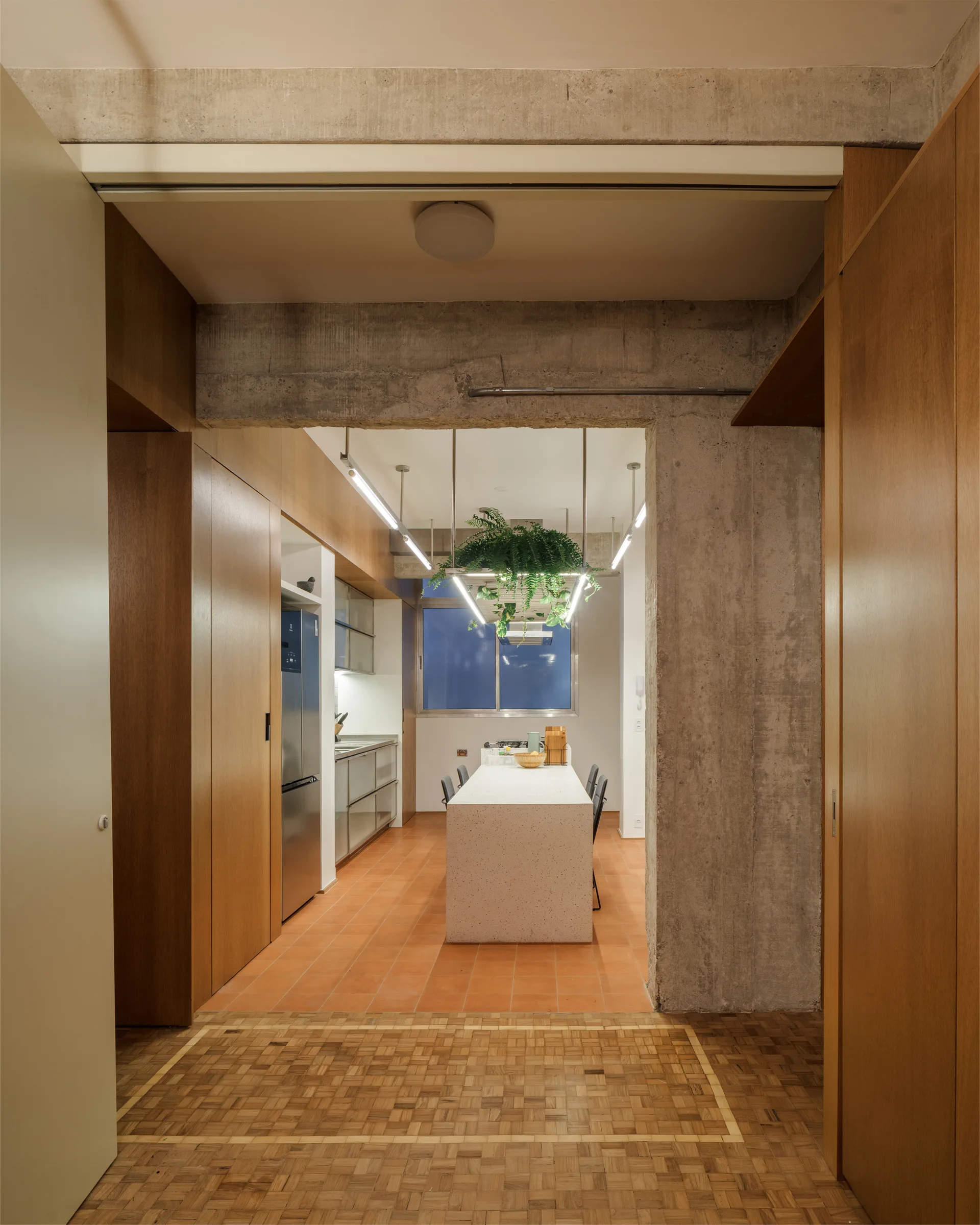 A view through a concrete archway into a modern kitchen with wood cabinets, a speckled stone island, linear light fixture, and terracotta floor tiles.