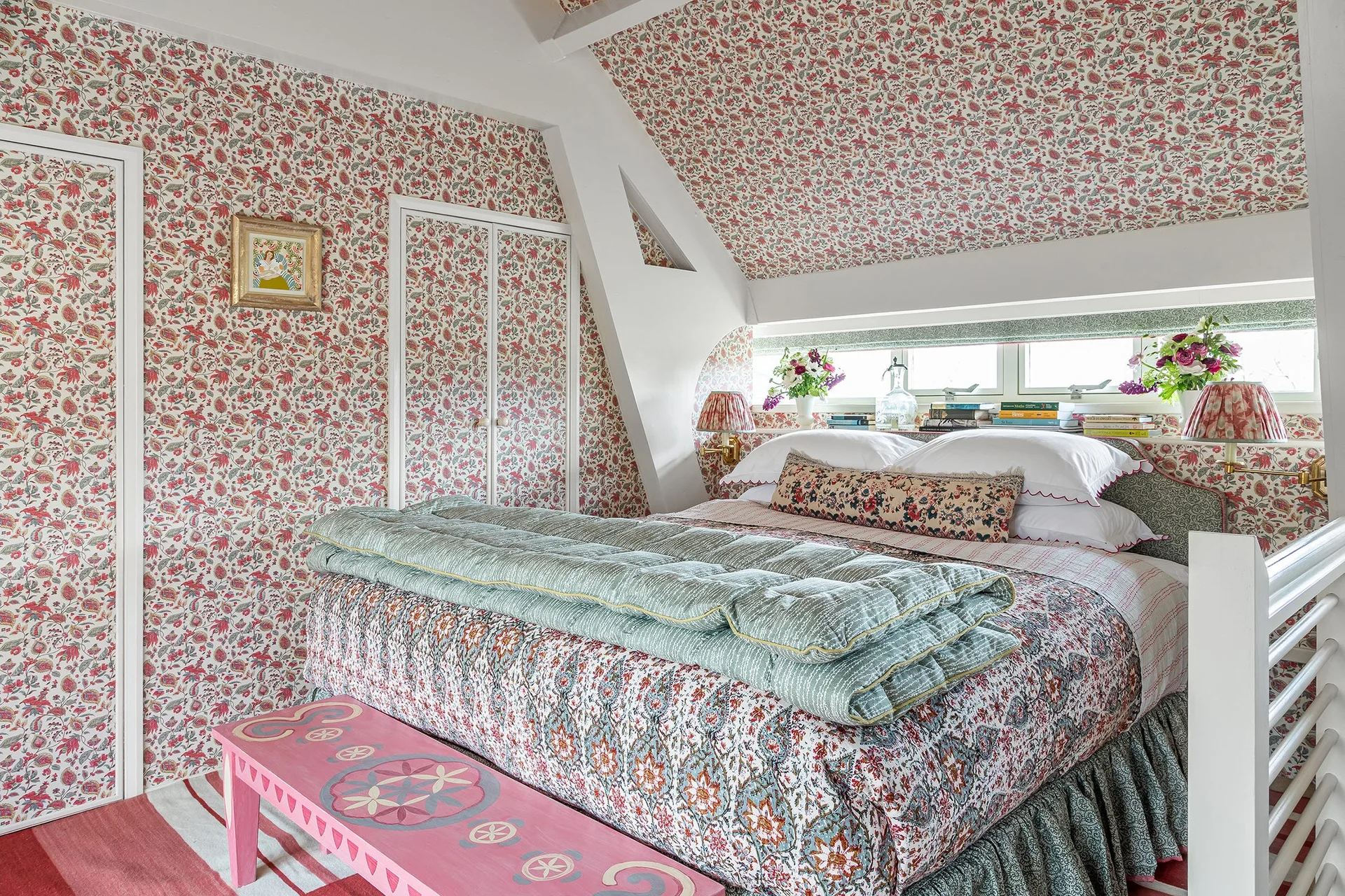 An attic bedroom with walls and a sloped ceiling covered in pink floral wallpaper, featuring a bed with layered quilts and a pink bench.