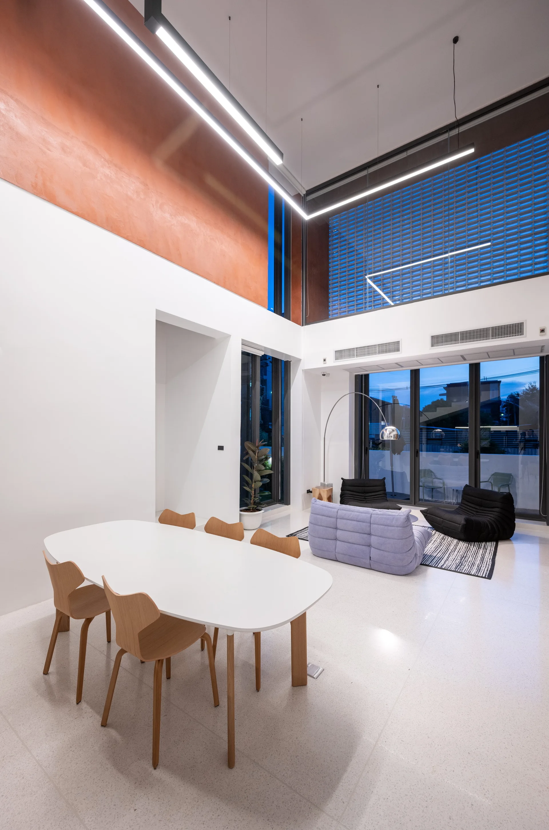 A contemporary open-plan dining and living area features an oval table, light wood chairs, a lavender modular sofa, and a high terracotta wall accent.