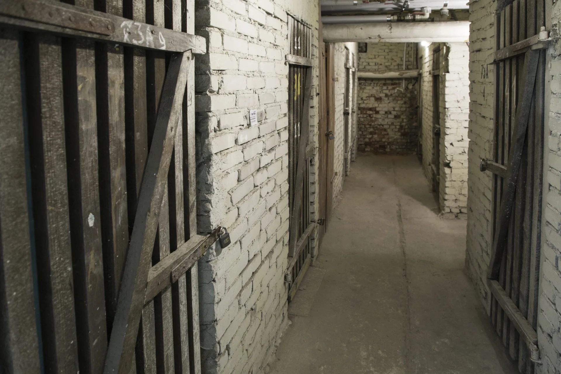 A narrow basement hallway with painted white brick walls, concrete flooring, and rustic wooden slatted storage doors.