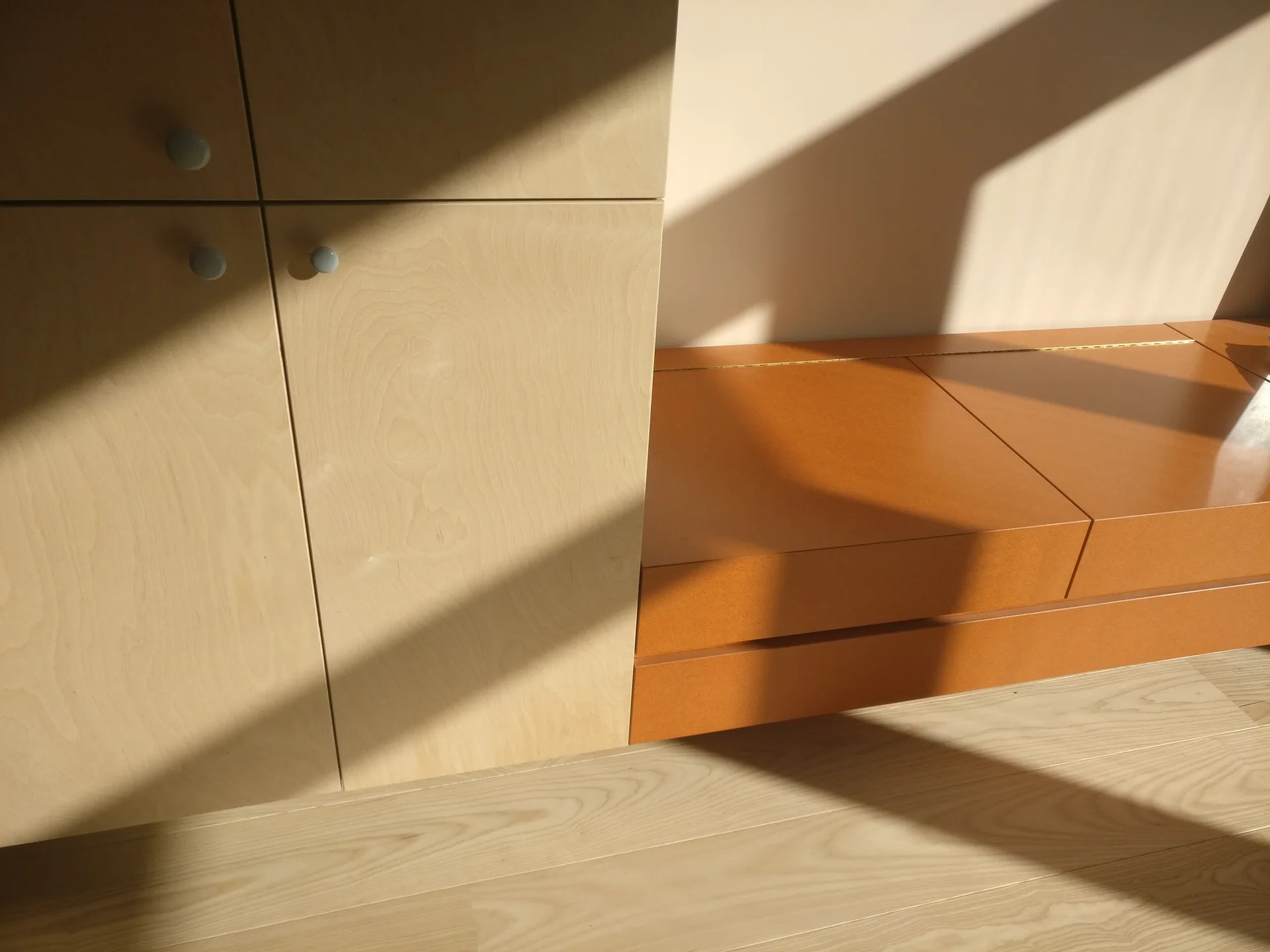 A corner view of light wood cabinetry and a warm orange storage bench on hardwood floors, highlighted by sharp geometric shadows from sunlight.