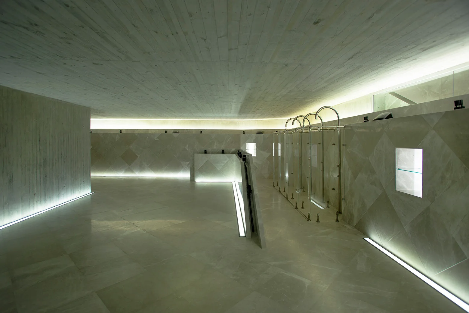 A spacious minimalist wellness room featuring glass shower stalls, diamond-patterned stone walls, and recessed linear lighting along the floor.