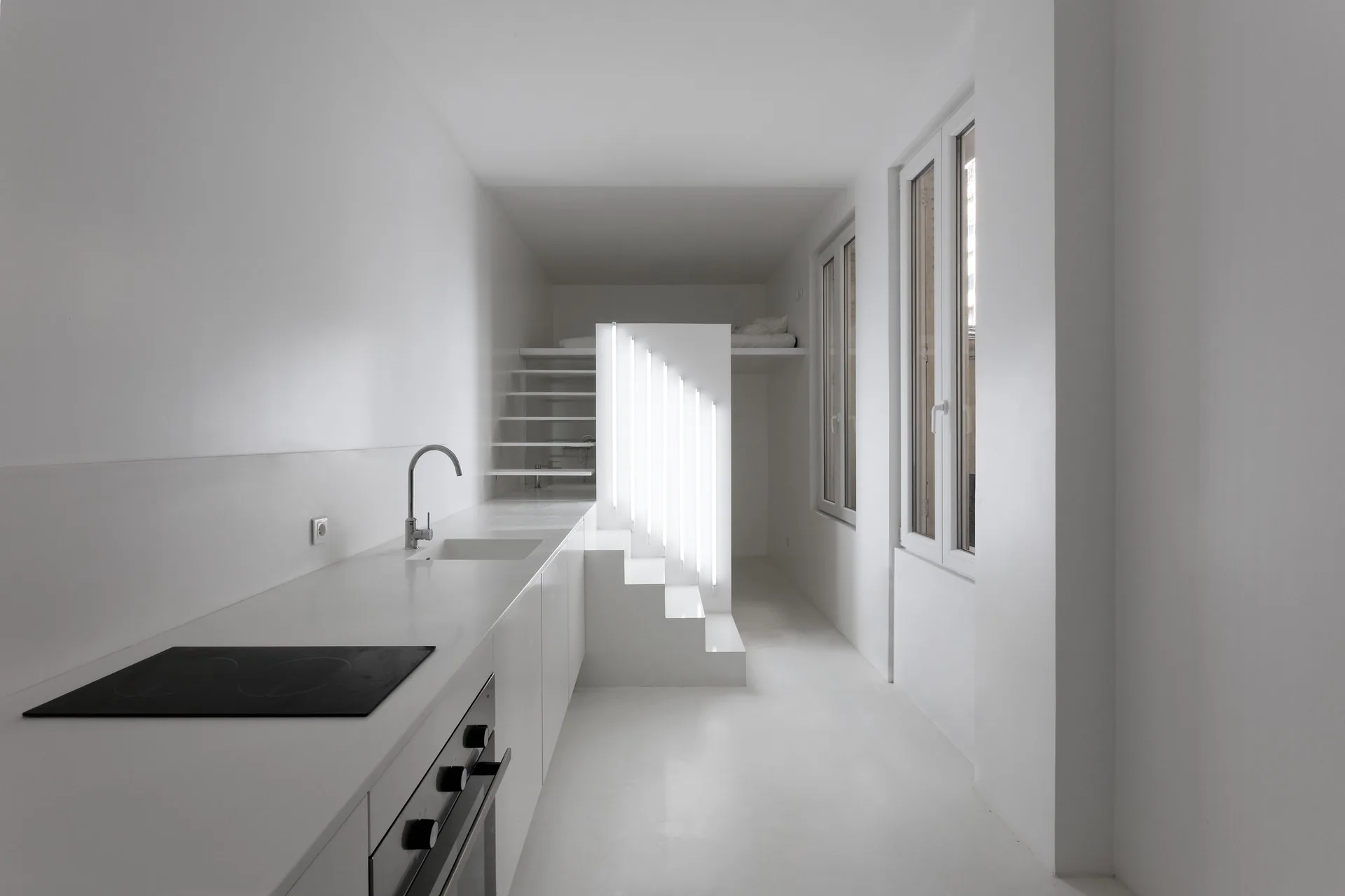 A monochromatic white minimalist kitchen featuring integrated appliances, tall windows, and a staircase highlighted by vertical tube lights.