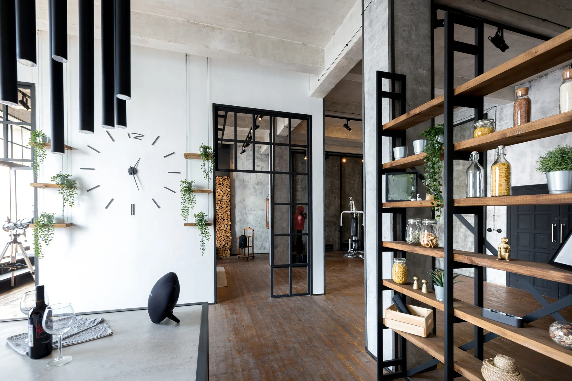 An industrial living room featuring a black metal grid partition, open wooden shelving, concrete ceilings, and a minimalist wall clock.