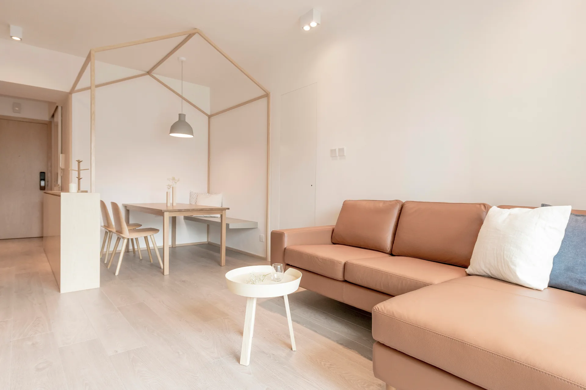 A minimalist open-concept living room features a tan leather sofa, a round white coffee table, and a wooden house-shaped frame over the dining table.