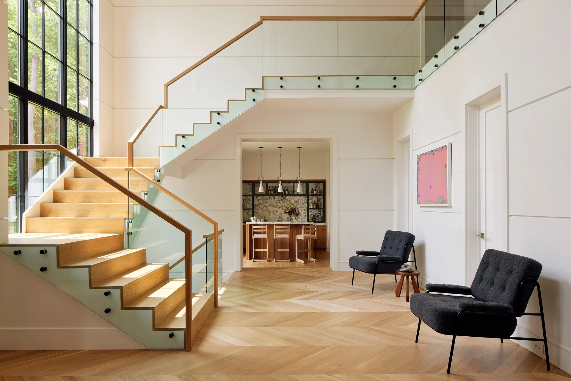 A spacious entryway features a light wood staircase with glass railings, chevron patterned wood flooring, and a pair of dark grey armchairs.