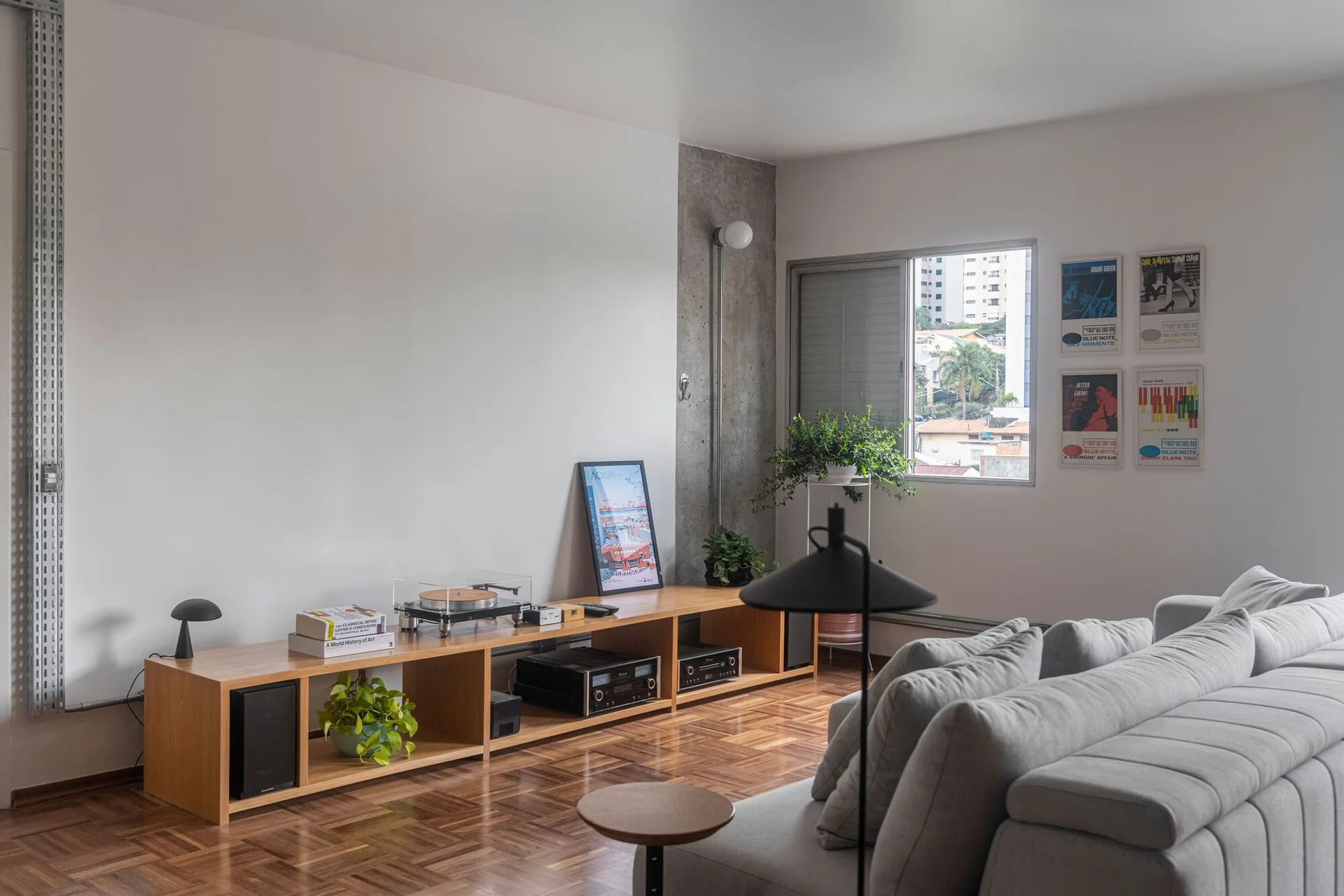 A minimalist living room features a low wooden media console, a grey sectional sofa, and distinctive herringbone parquet flooring under natural light.
