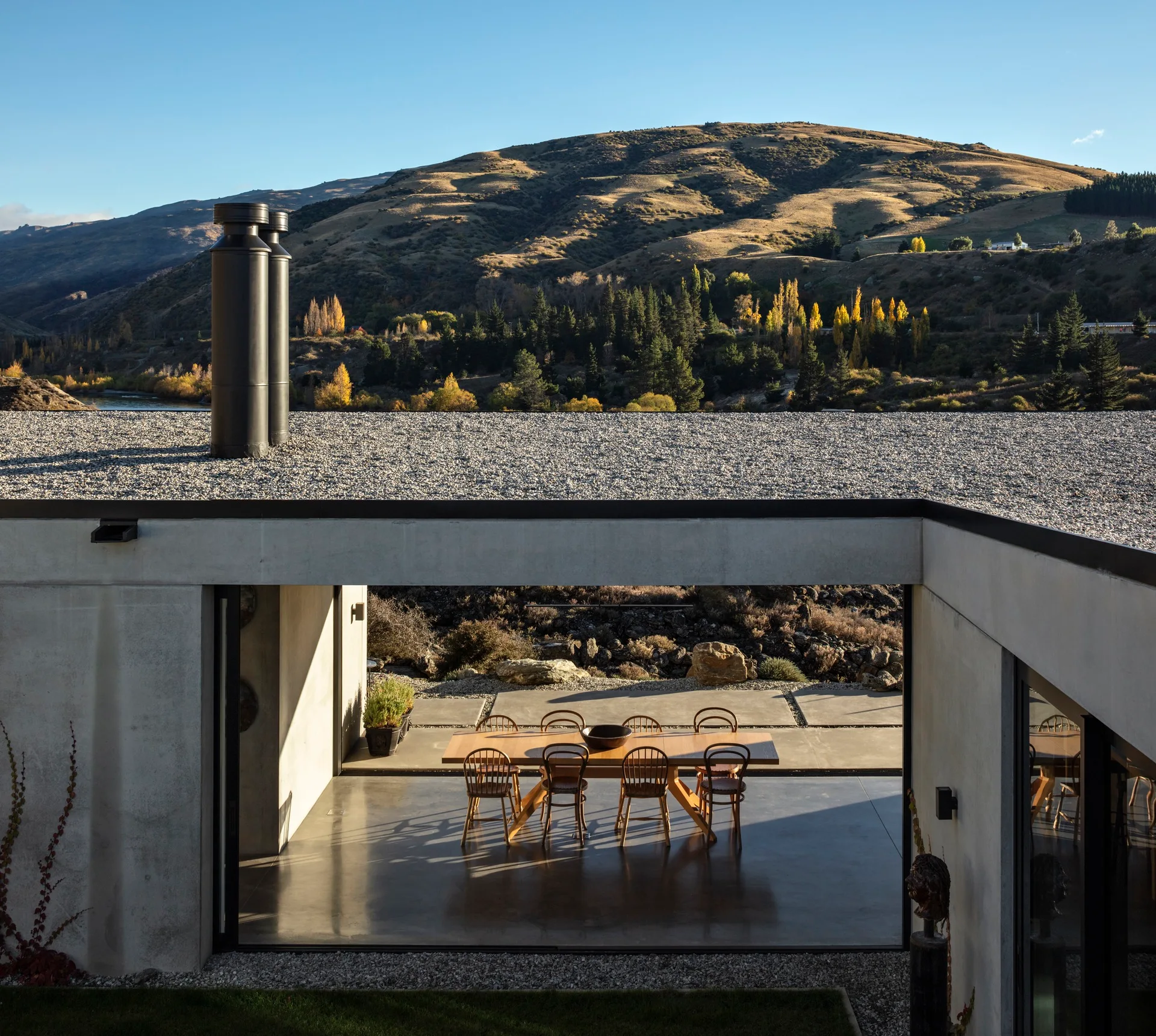 A wooden dining table and chairs sit on a concrete covered patio overlooking a mountainous landscape under a blue sky.