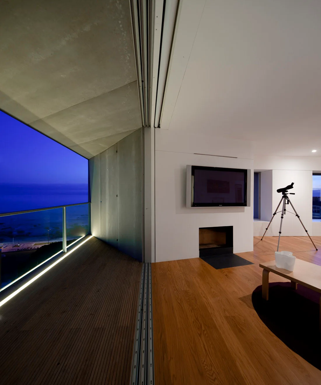 A minimalist living room with wood floors and a fireplace opens to a concrete balcony overlooking a dark blue twilight horizon.