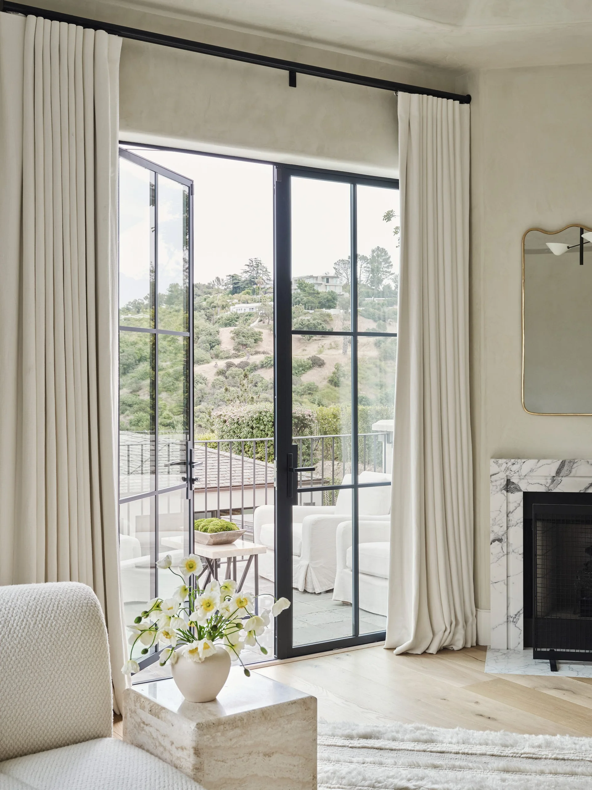 Black metal French doors open to a balcony, flanked by cream drapery, next to a marble fireplace and travertine side table.