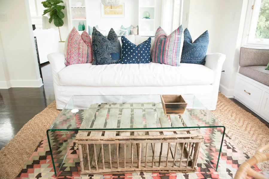A white slipcovered sofa with patterned blue and red pillows sits behind a glass coffee table with a rustic wood crate base.