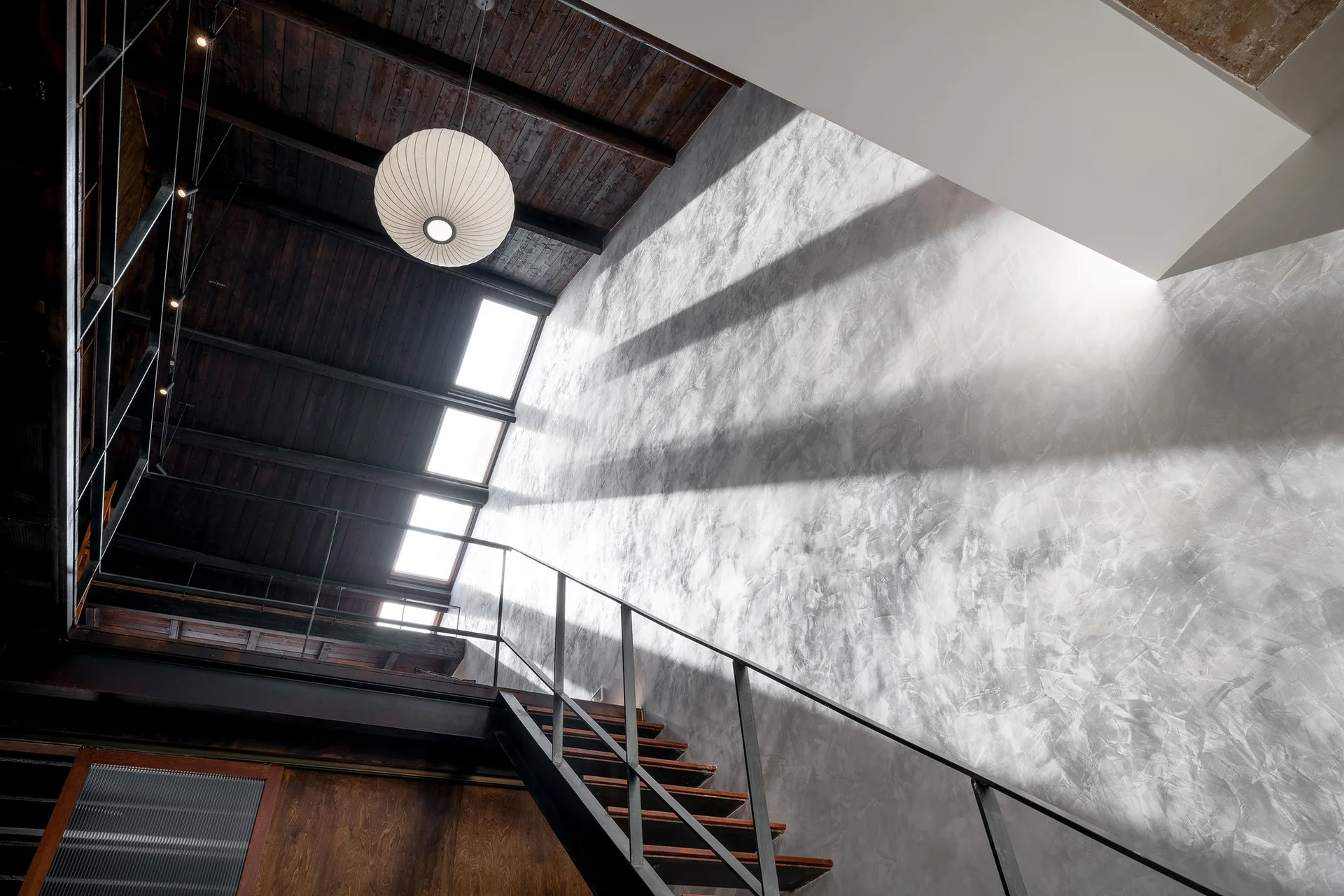 An industrial staircase with a metal railing rises next to a textured grey plaster wall under a dark wood ceiling with skylights.