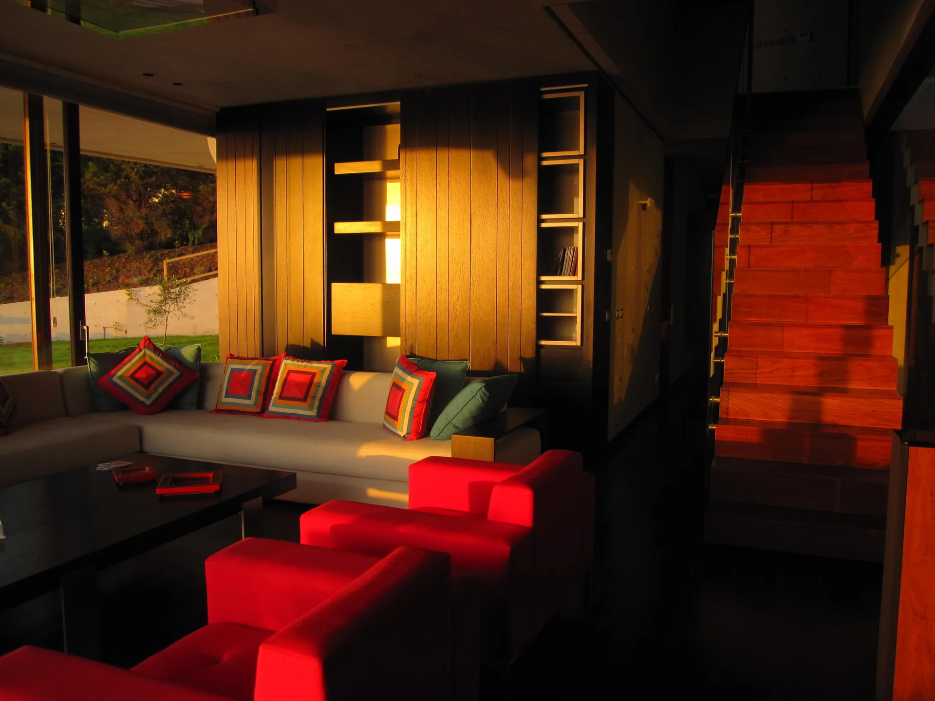 A modern living room features a light grey sectional sofa with geometric throw pillows, two bright red armchairs, and a wooden staircase.
