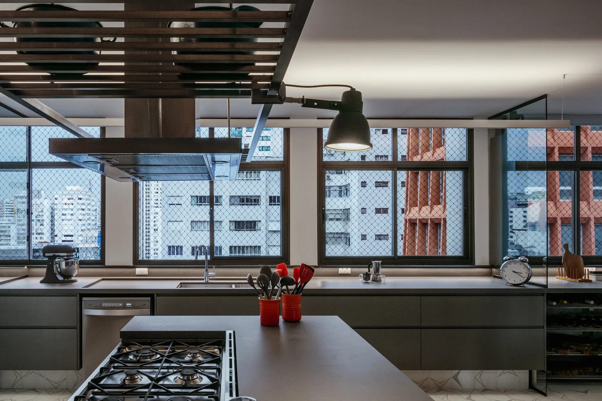 A modern kitchen with dark grey cabinetry, an island with a gas cooktop, a metal ceiling rack, and large windows with grid patterns.