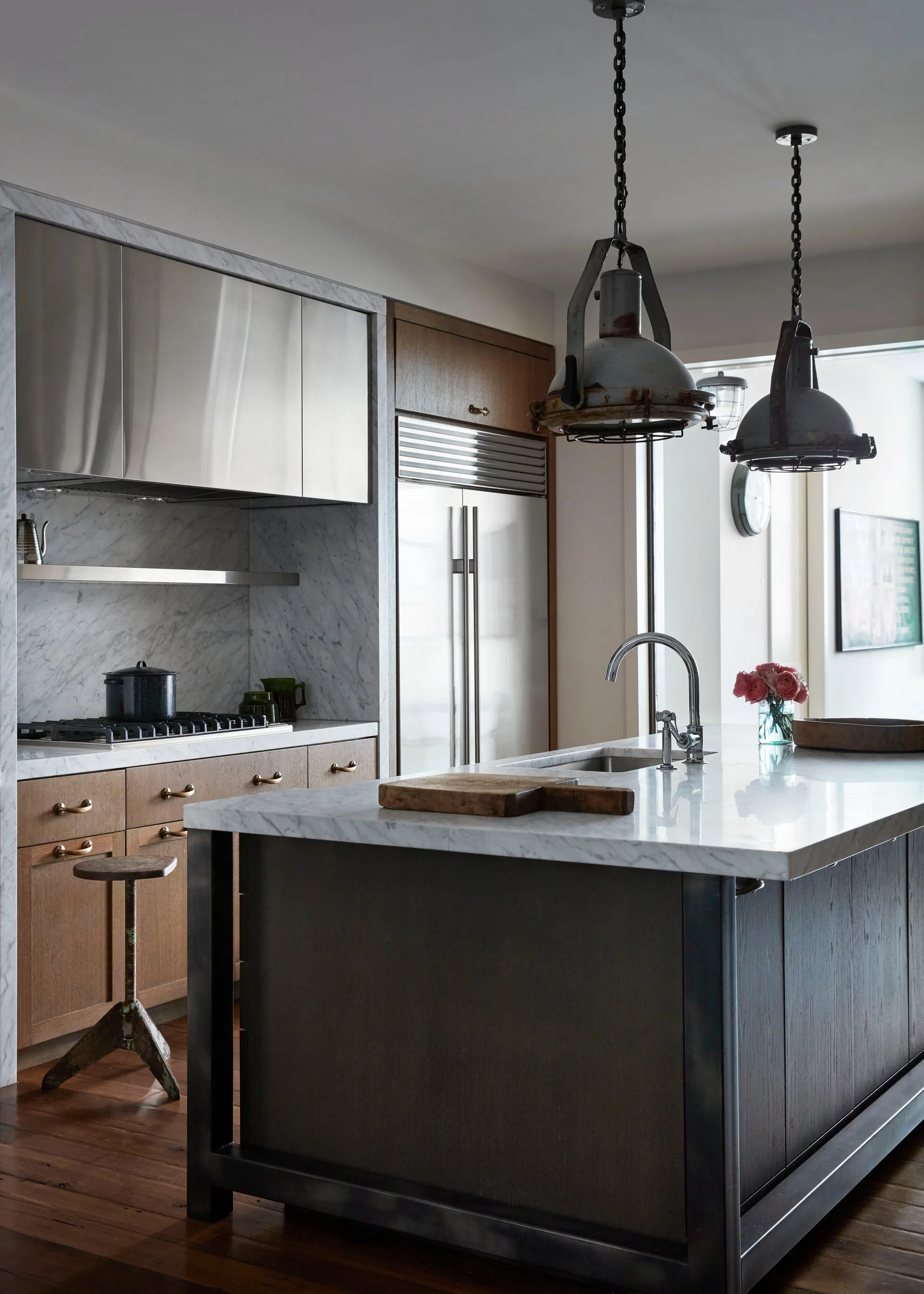A modern kitchen featuring a marble-topped island, wood cabinetry, stainless steel appliances, and two hanging industrial pendant lights.