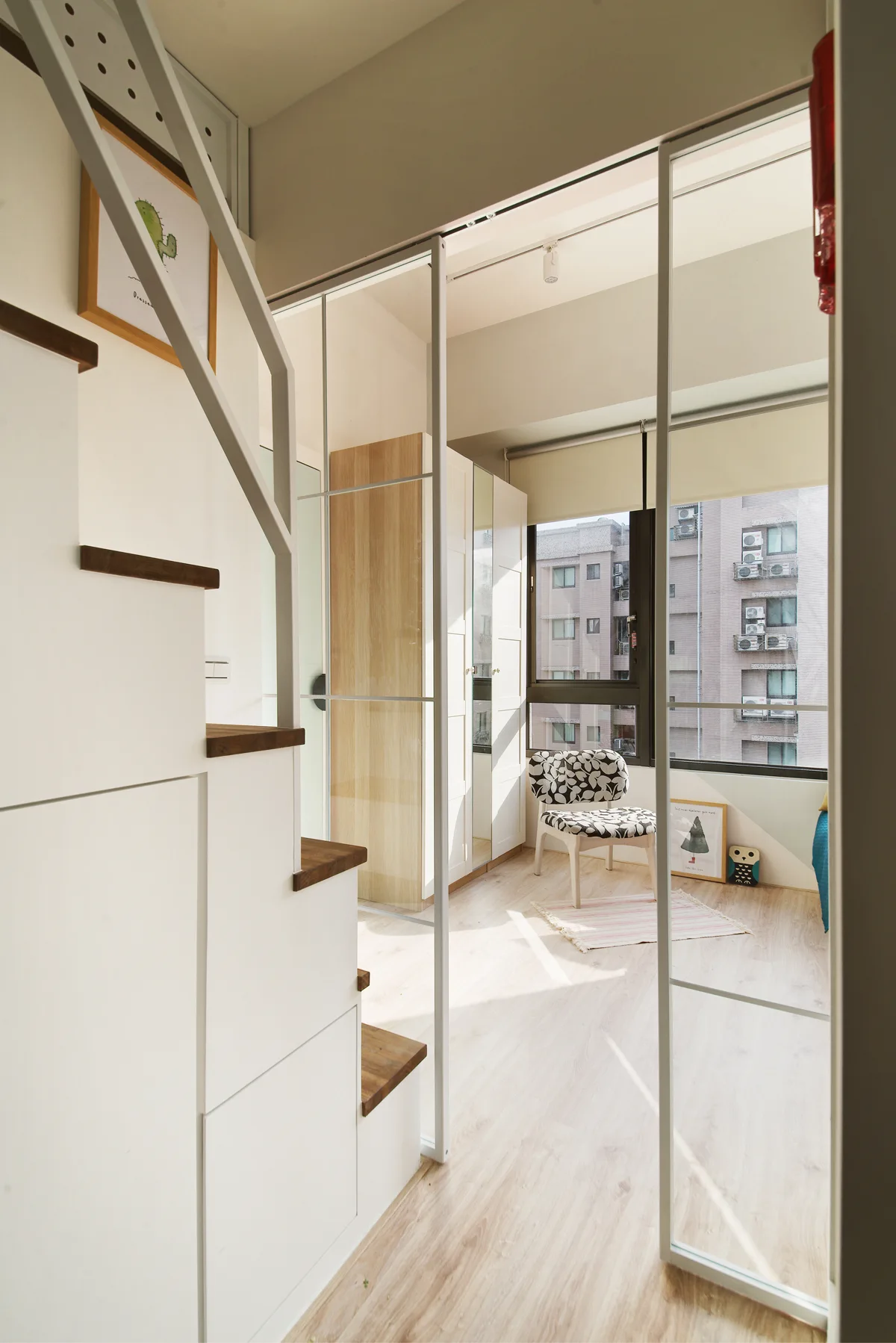 A compact loft bedroom with wood-tread stairs containing built-in storage drawers next to a white sliding glass partition.
