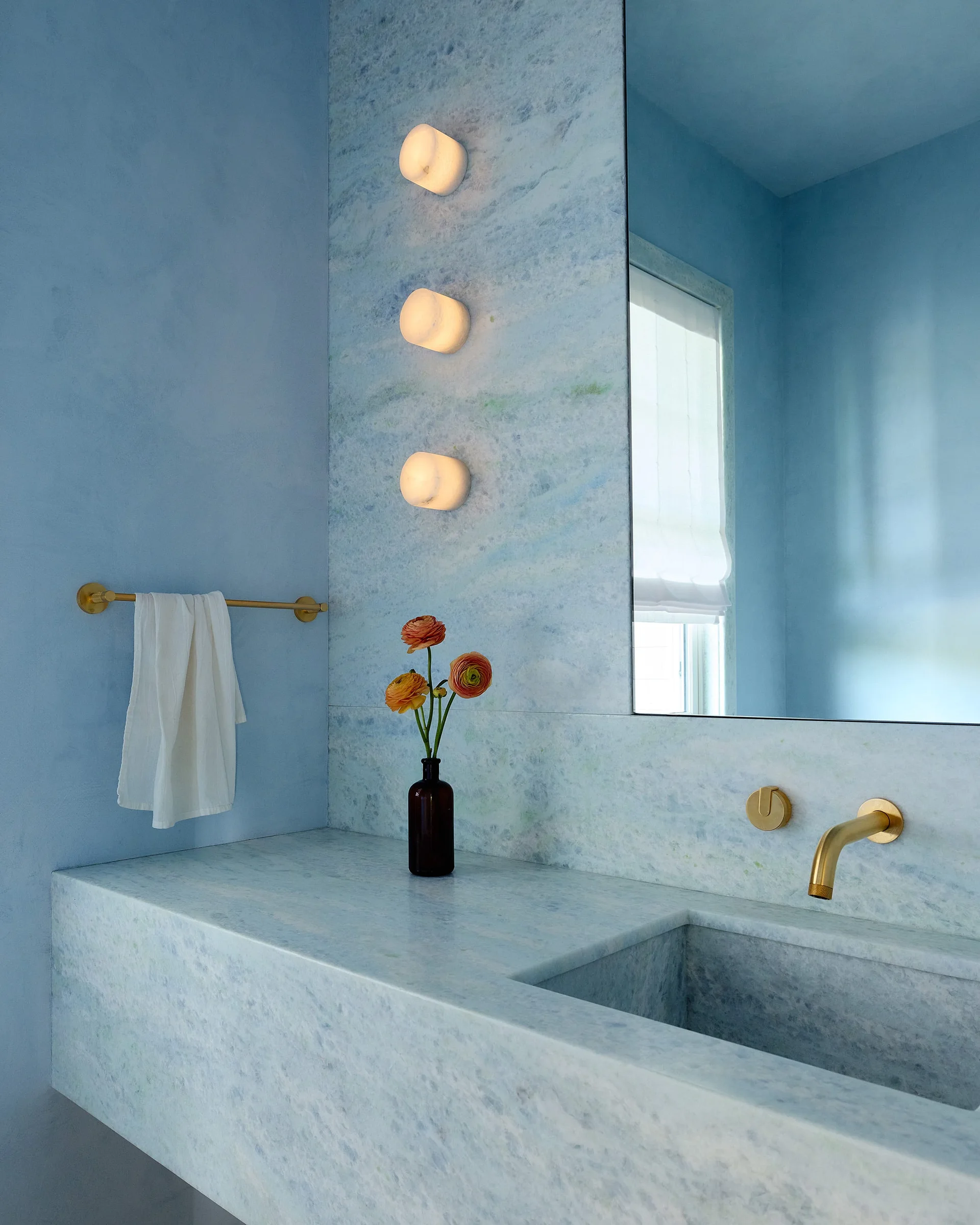 A bathroom vanity features a blue veined stone countertop, an integrated sink with a wall-mounted brass faucet, and three vertically stacked sconces.
