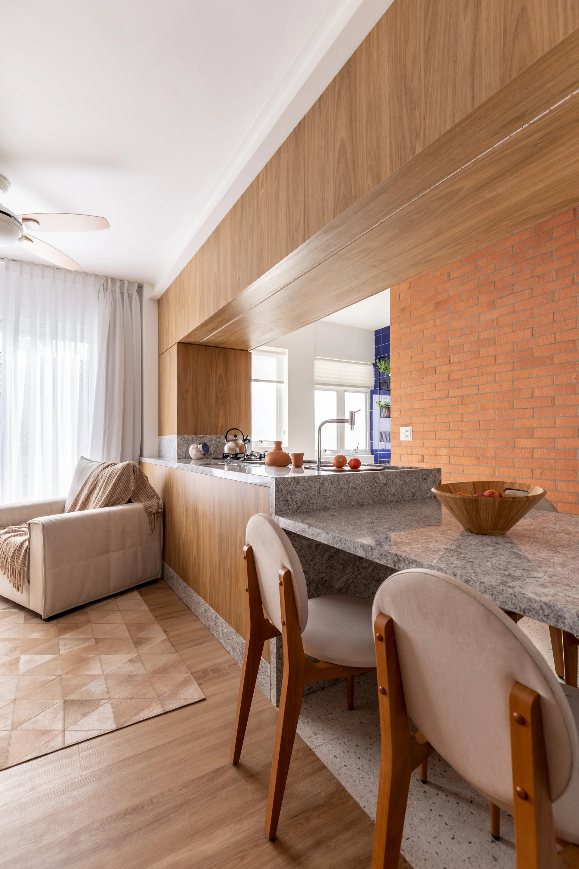 A modern kitchen featuring a wood and stone island with a lowered dining counter, upholstered wooden chairs, and an exposed brick accent wall.
