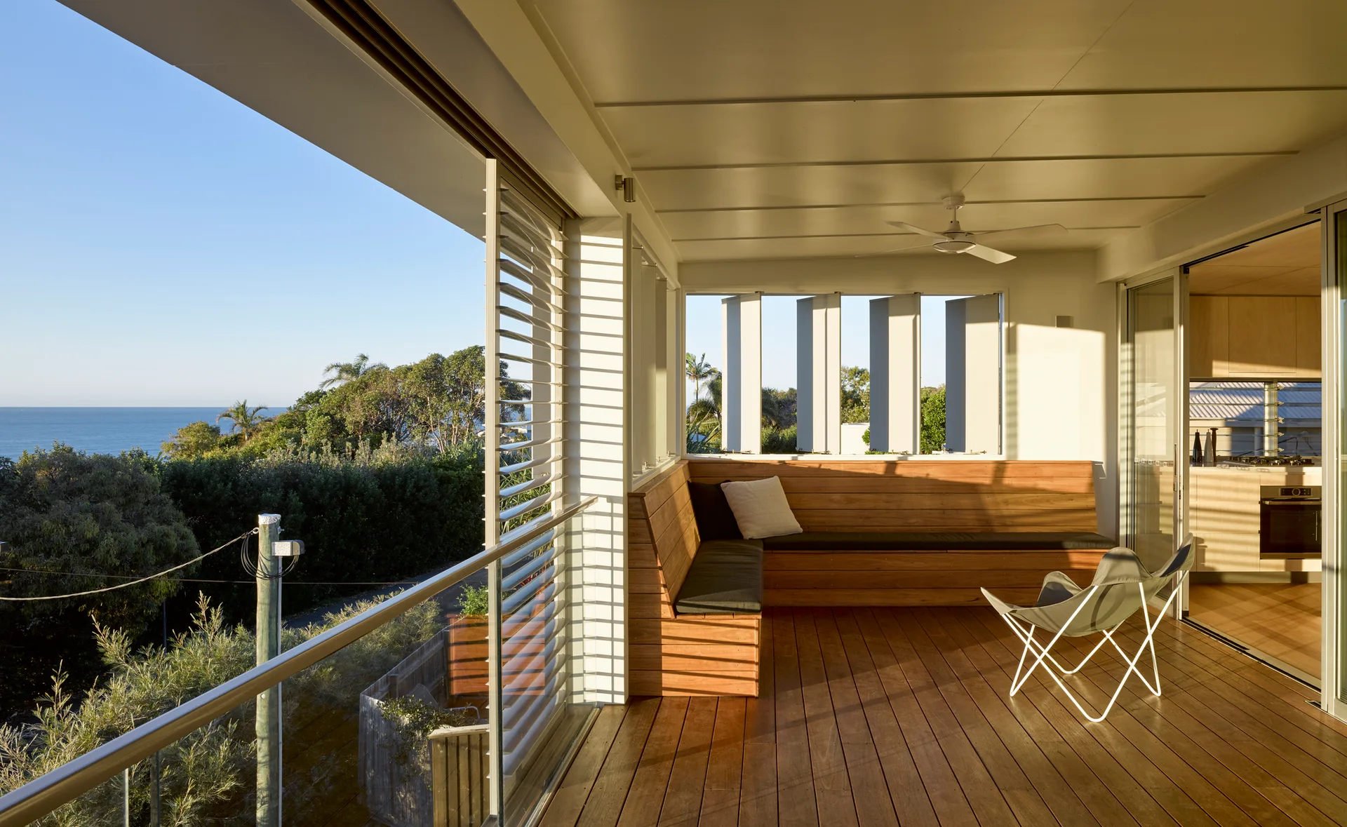 A covered wooden balcony features an integrated timber corner bench, a modern folding lounge chair, and a glass railing overlooking the ocean.