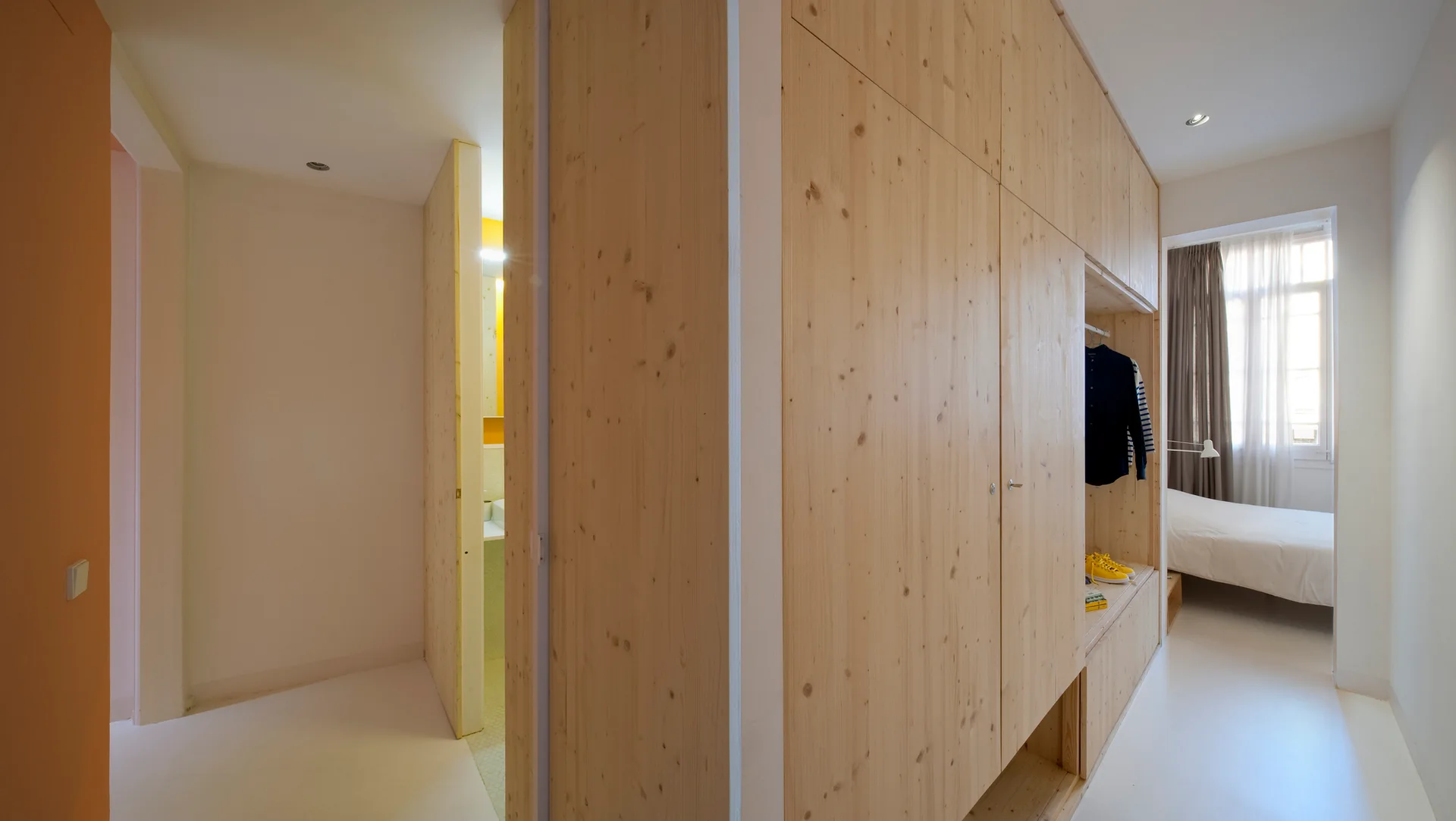 A minimalist white bedroom featuring a large custom light wood wardrobe unit that serves as a room divider next to the bathroom entry.