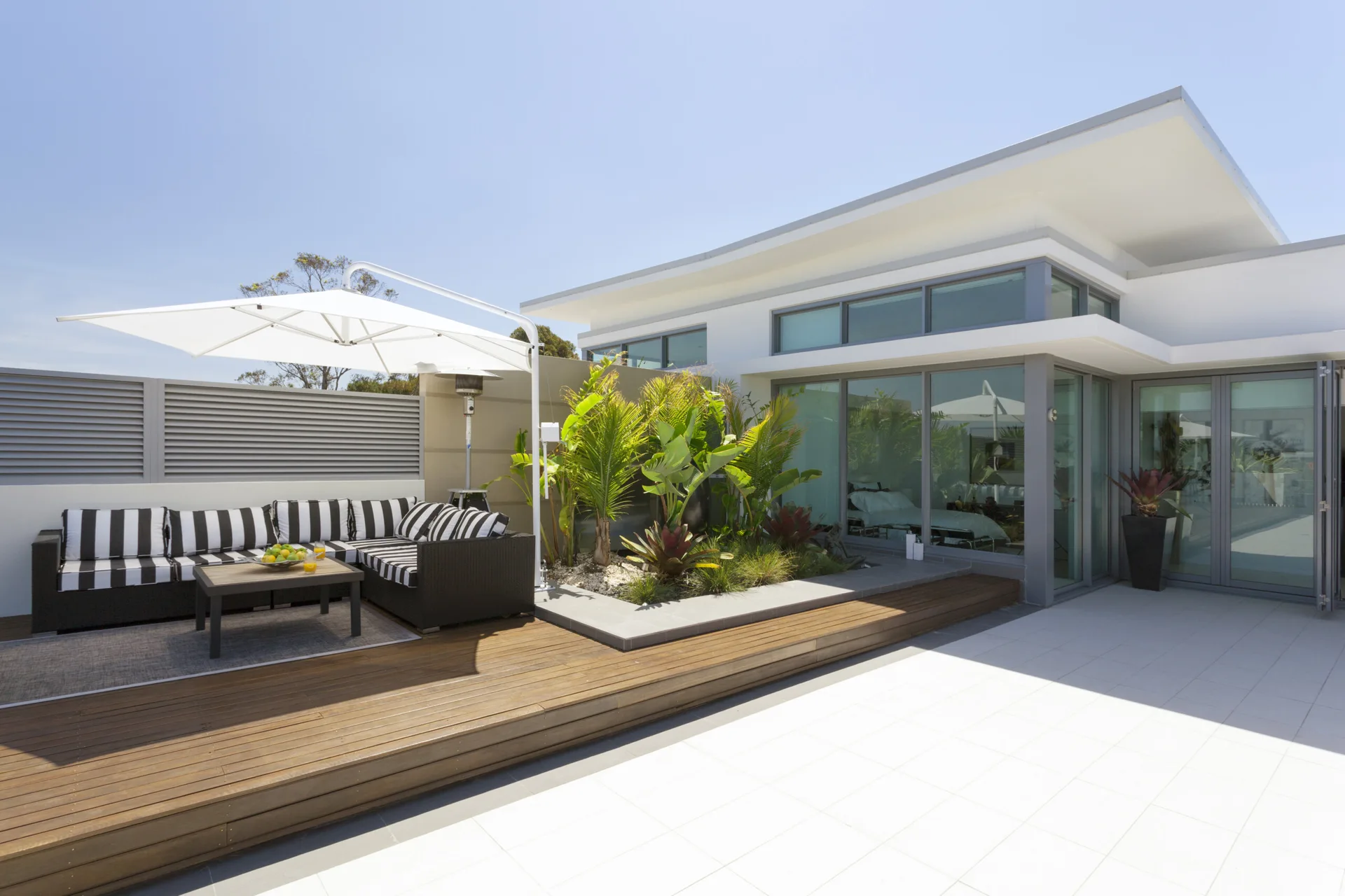 A modern wood deck features a black and white striped sectional sofa under a white cantilever umbrella next to a raised tropical garden bed.