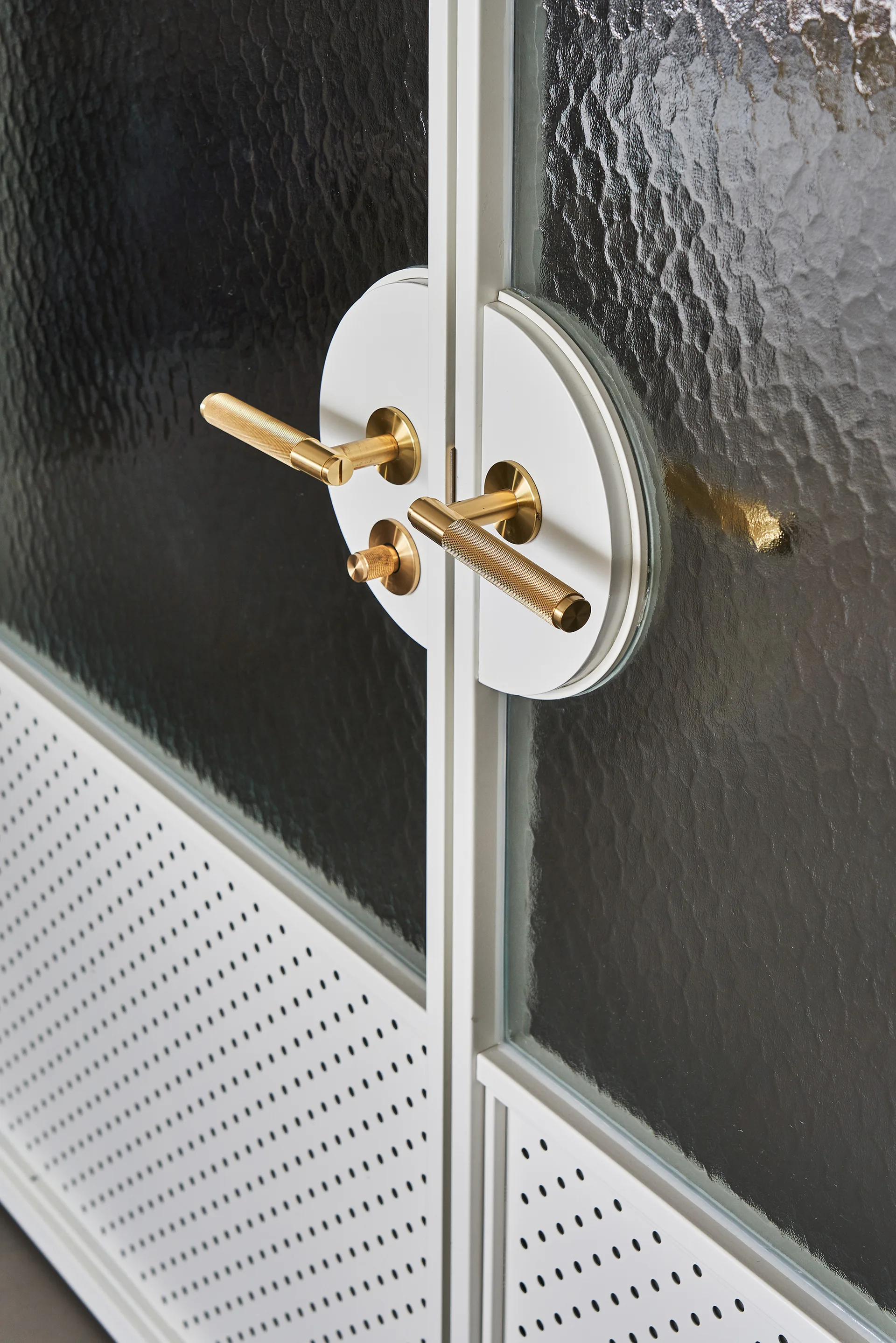 Close-up of white metal double doors with textured glass panels, perforated lower panels, and knurled brass lever handles.