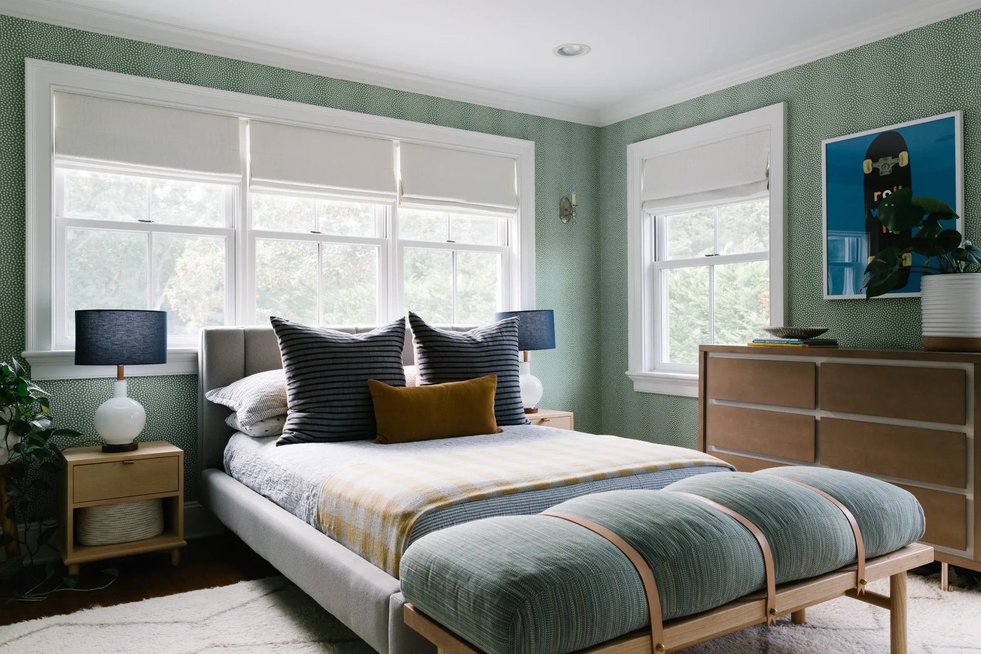 A bedroom features green patterned wallpaper, a gray upholstered bed with striped pillows, two light wood nightstands, and a cushioned bench at the foot.