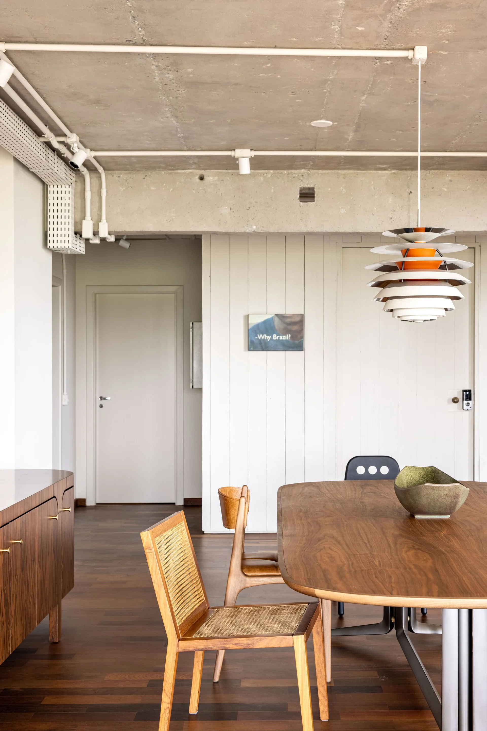 A wooden dining table and cane chairs sit under an exposed concrete ceiling with a layered pendant light in a minimalist home.