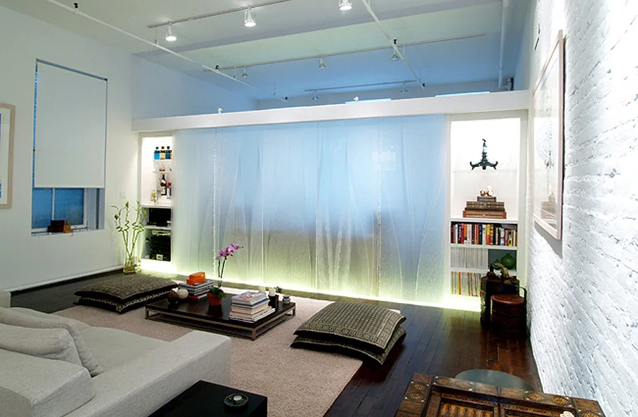 A modern living room with a low coffee table, floor cushions, an illuminated translucent screen divider, and white painted brick walls.