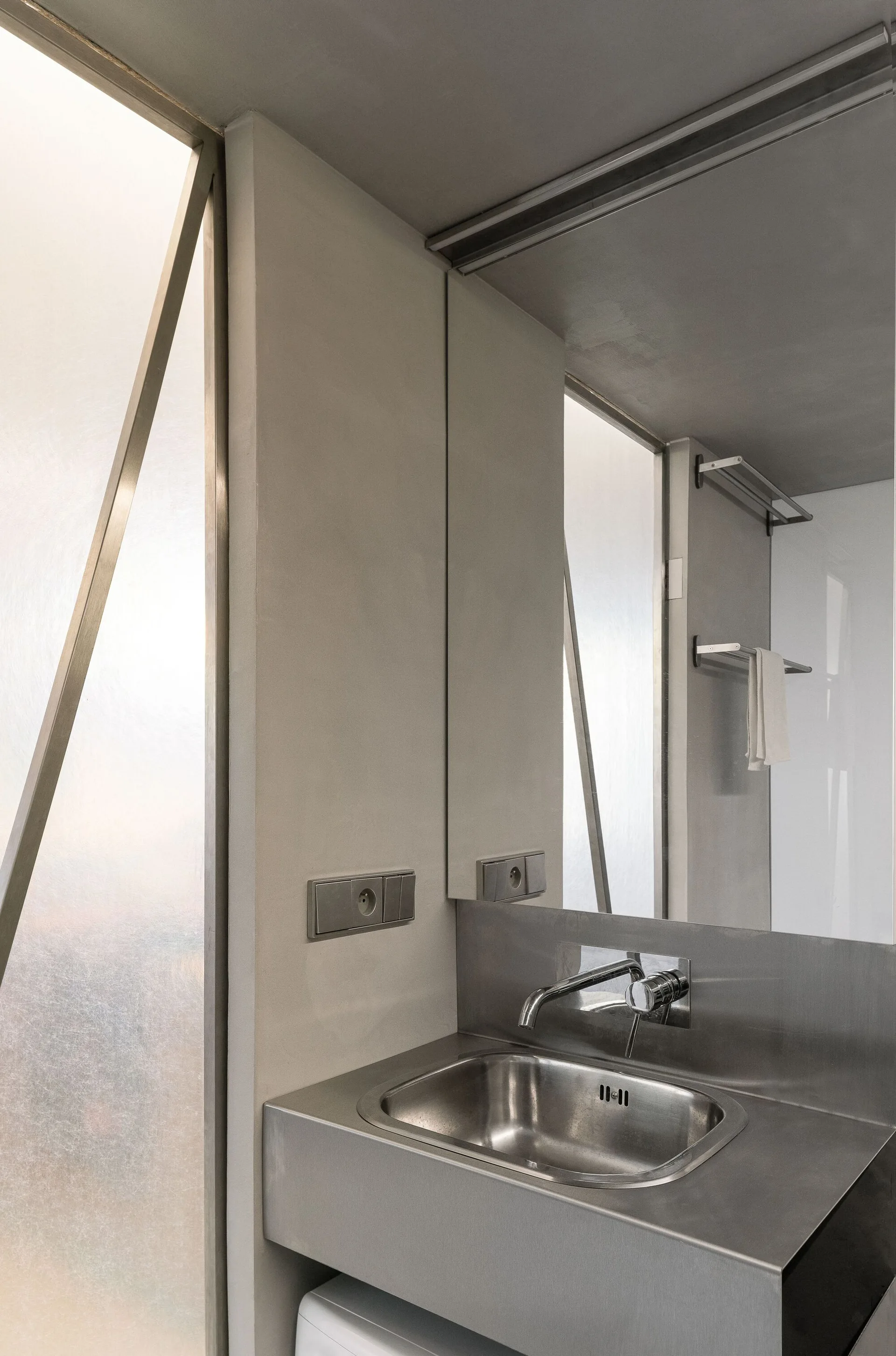 A minimalist bathroom vanity features a custom stainless steel sink and a frosted glass wall partition.