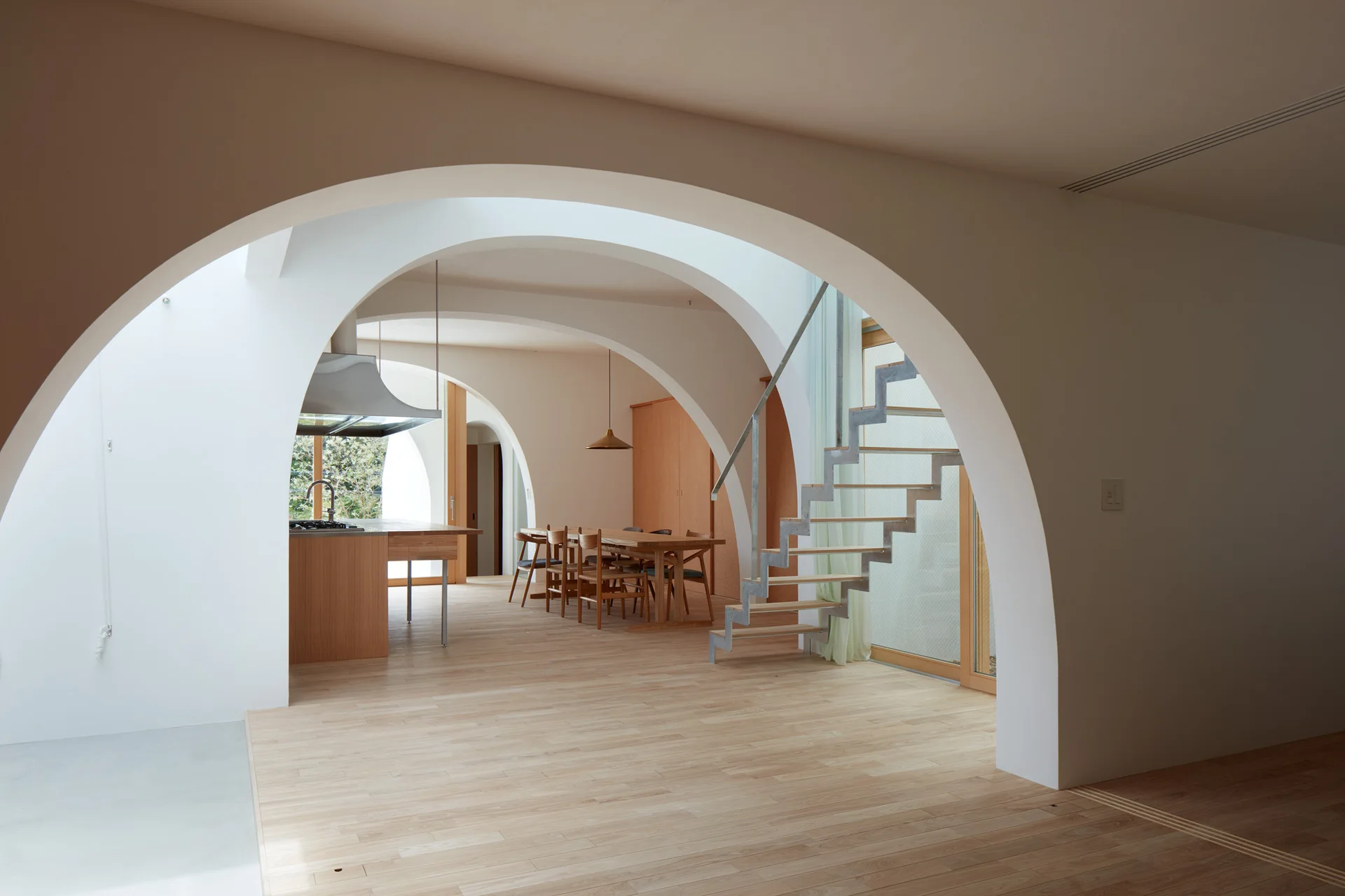 A minimalist open-plan home features multiple smooth white plaster archways, light wood floors, an open-riser staircase, and a dining table.
