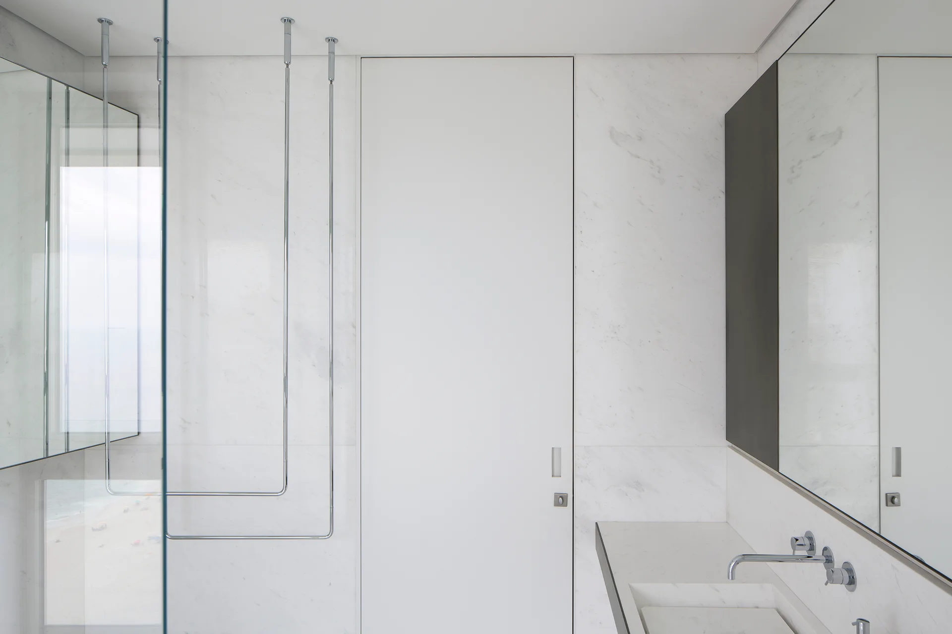 Minimalist white marble bathroom with a flush floor-to-ceiling door, integrated basin, and ceiling-mounted chrome towel racks.