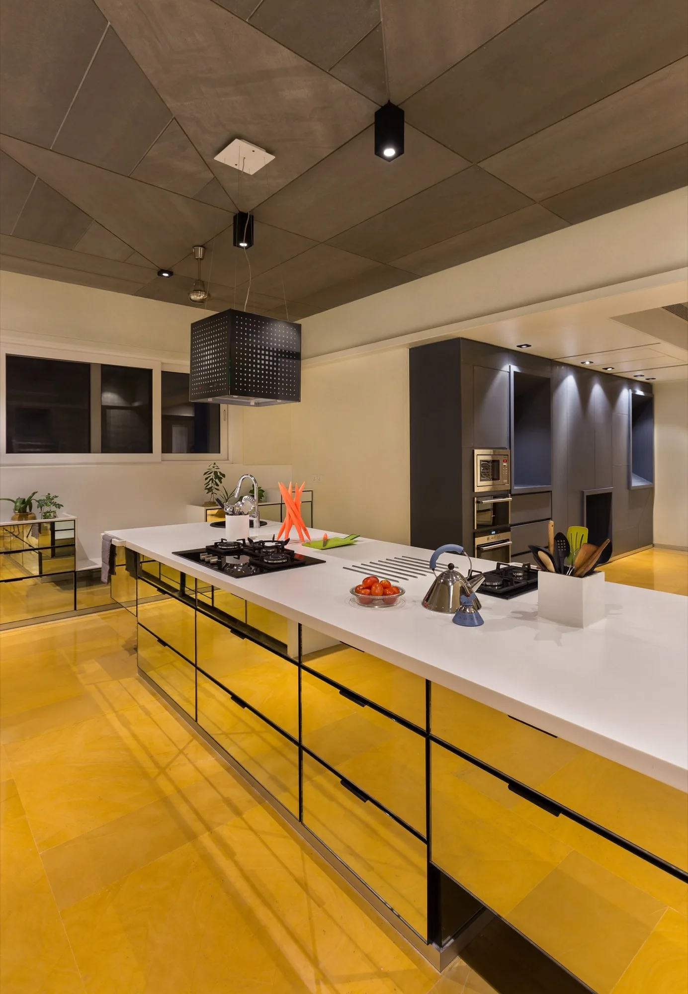 A modern kitchen with a white-countertop island, mirrored cabinets reflecting yellow tiled floors, and a grey geometric panel ceiling.