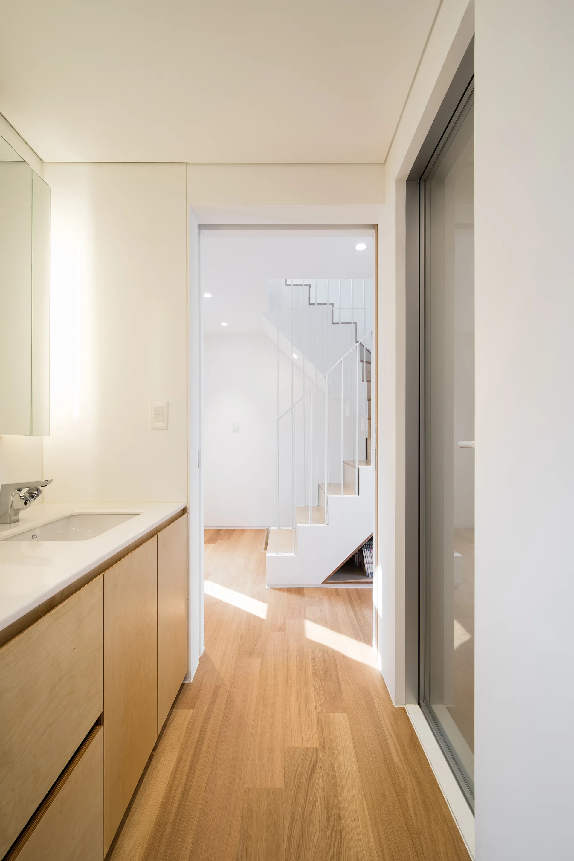 A minimalist bathroom with a light wood vanity and white sink leads into a bright hallway showing a white metal staircase.