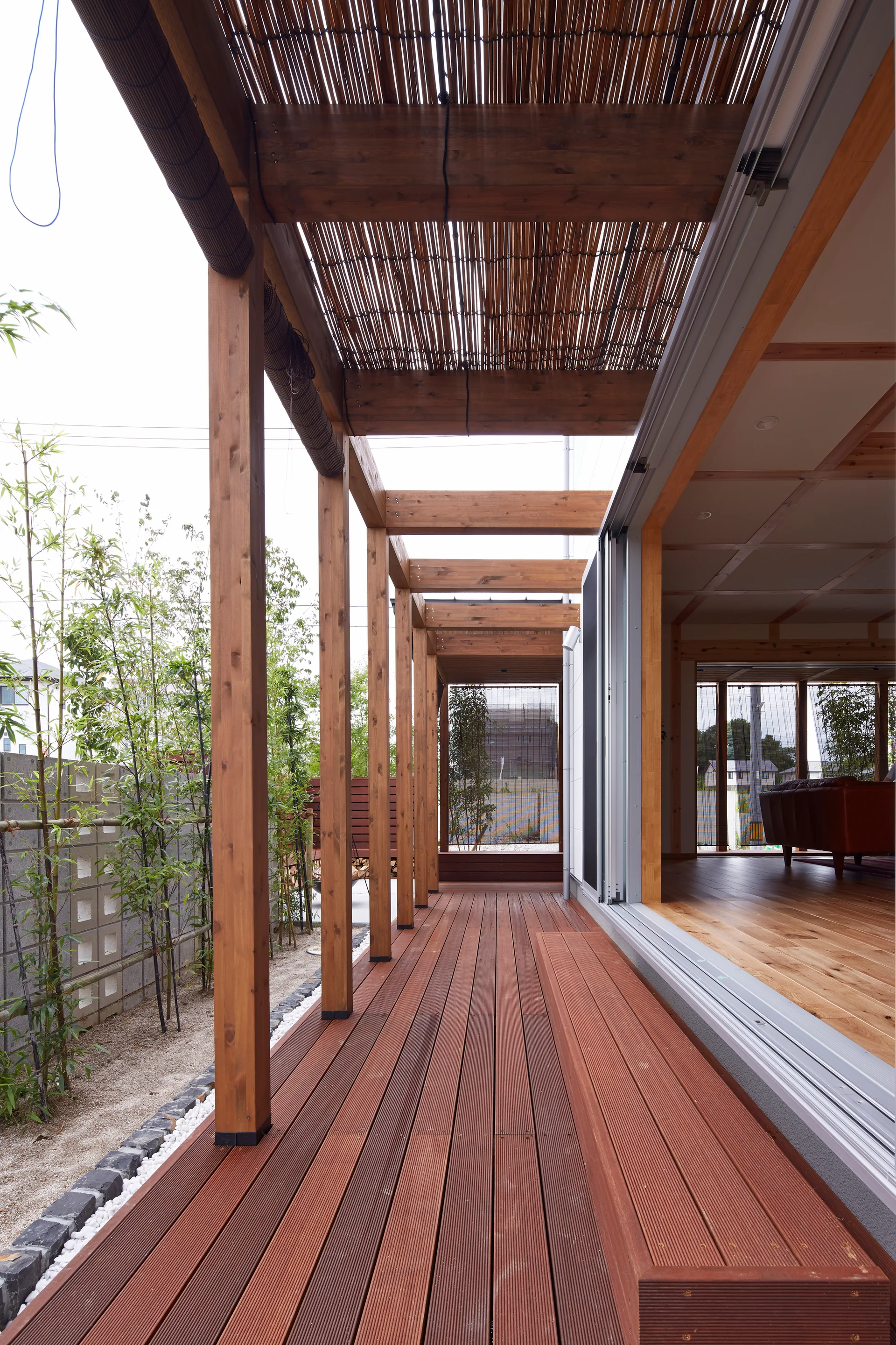 A long wooden deck with an overhead timber pergola runs alongside sliding glass doors that open to an indoor living room with bamboo landscaping outside.