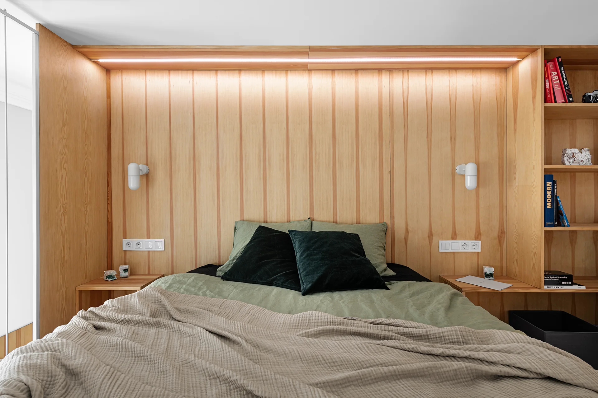 A bed with sage green linens sits against a vertical wood-paneled headboard with built-in side shelves and warm linear lighting.