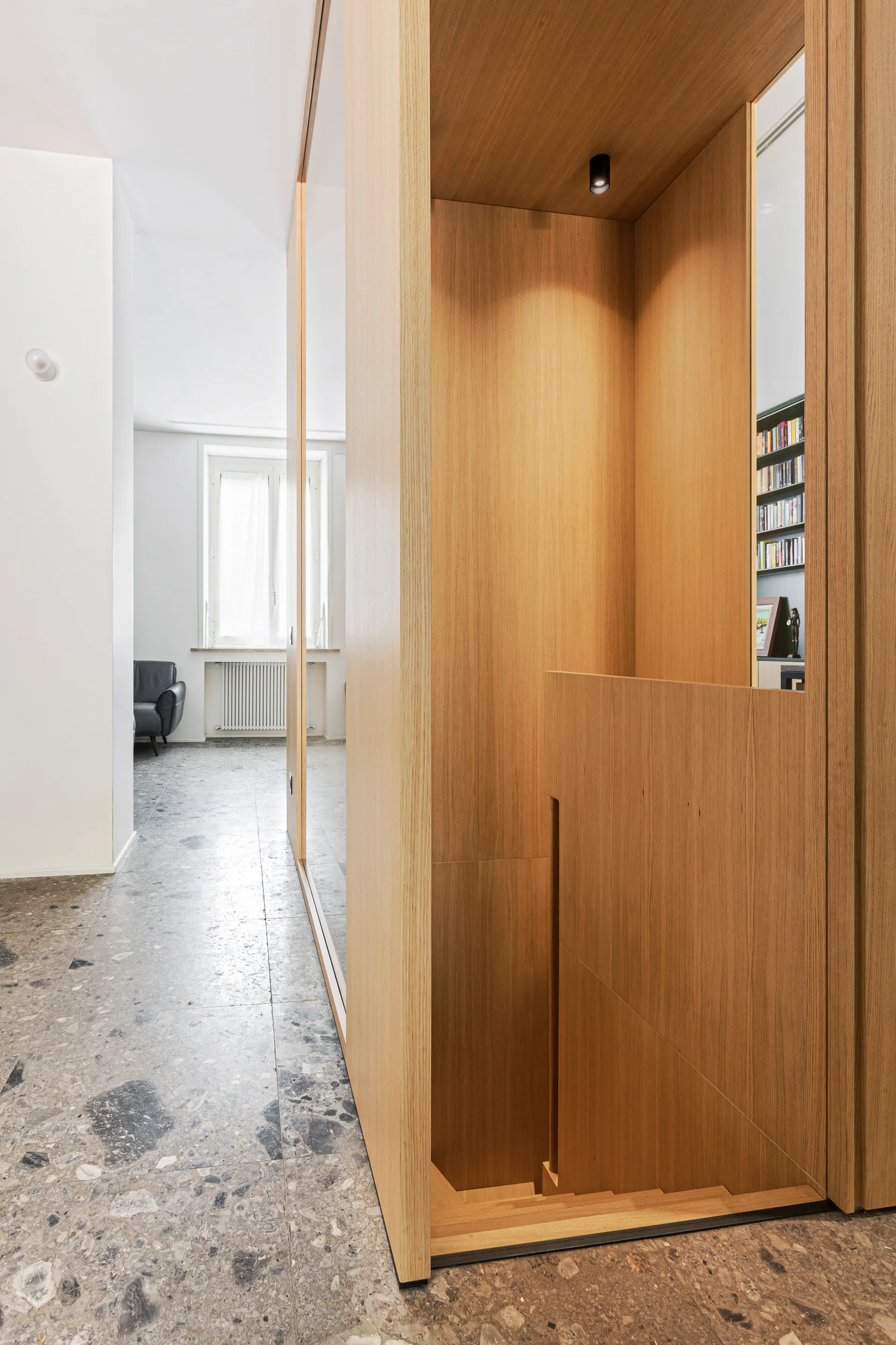 A light wood enclosed staircase descends from a polished grey terrazzo floor in a minimalist home interior.