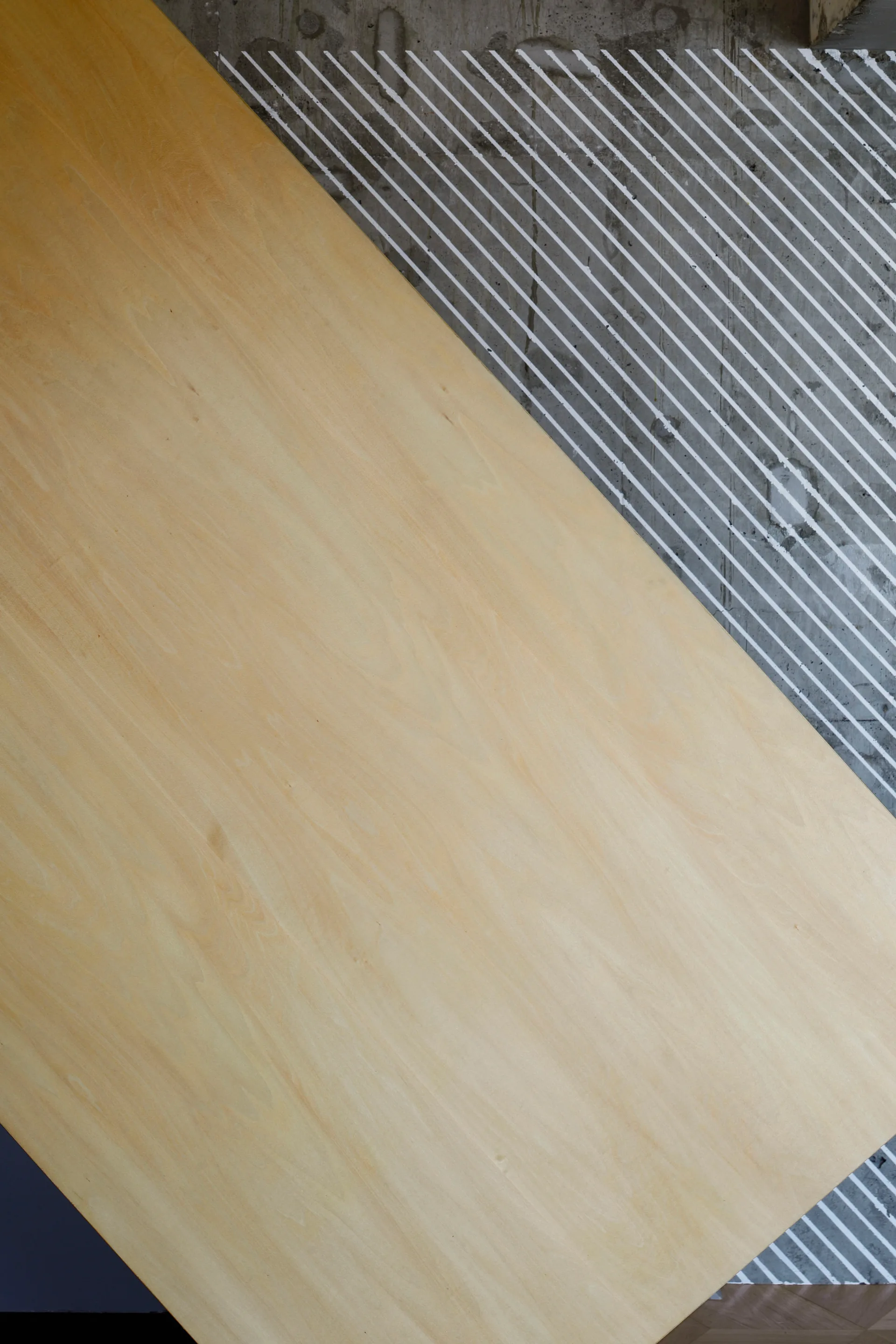 A light wood panel stands diagonally against a textured grey concrete wall accented with white painted diagonal stripes.