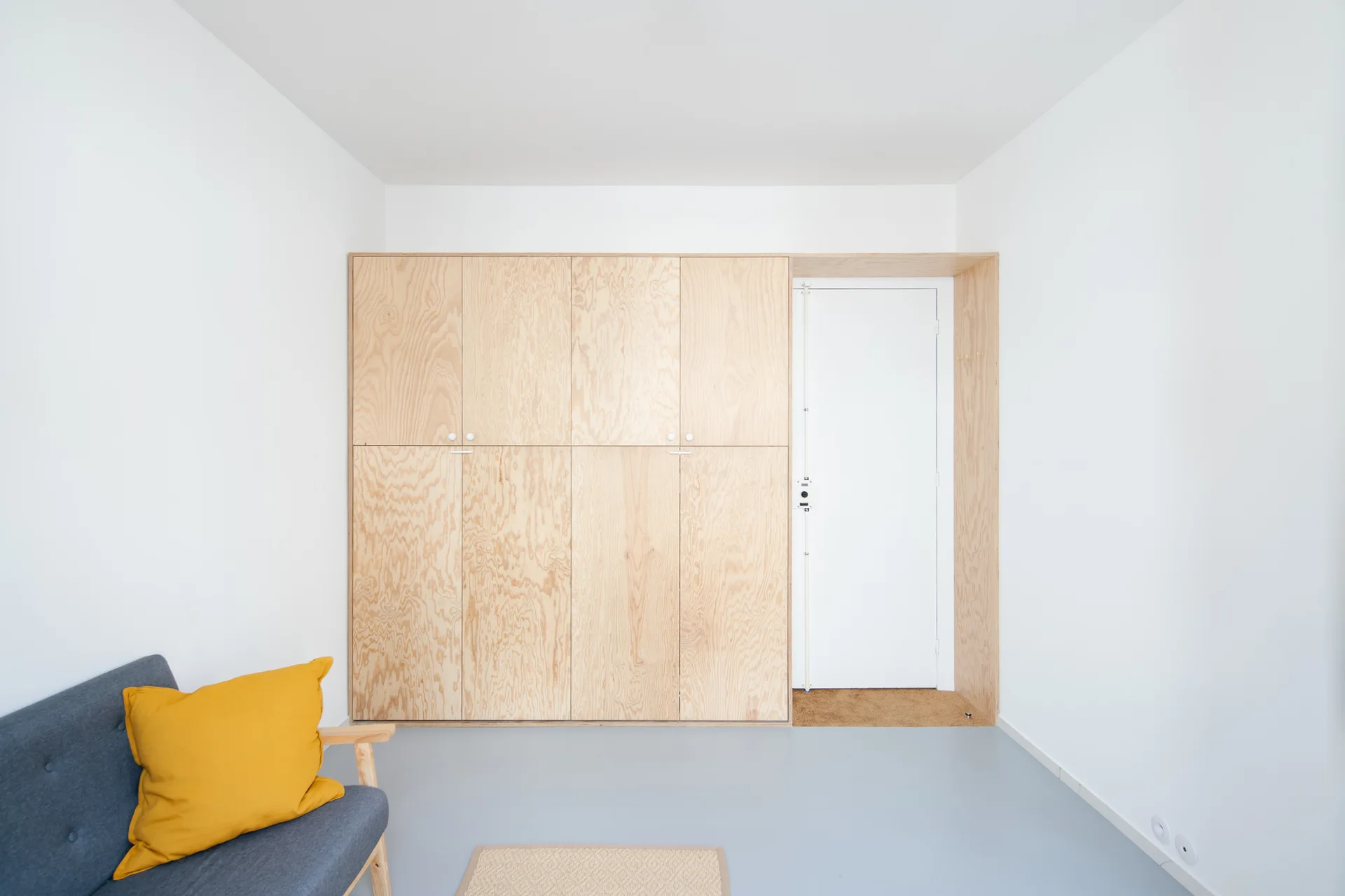 A minimalist entryway features a large built-in plywood wardrobe next to a white door, with a grey sofa and yellow pillow in the foreground.