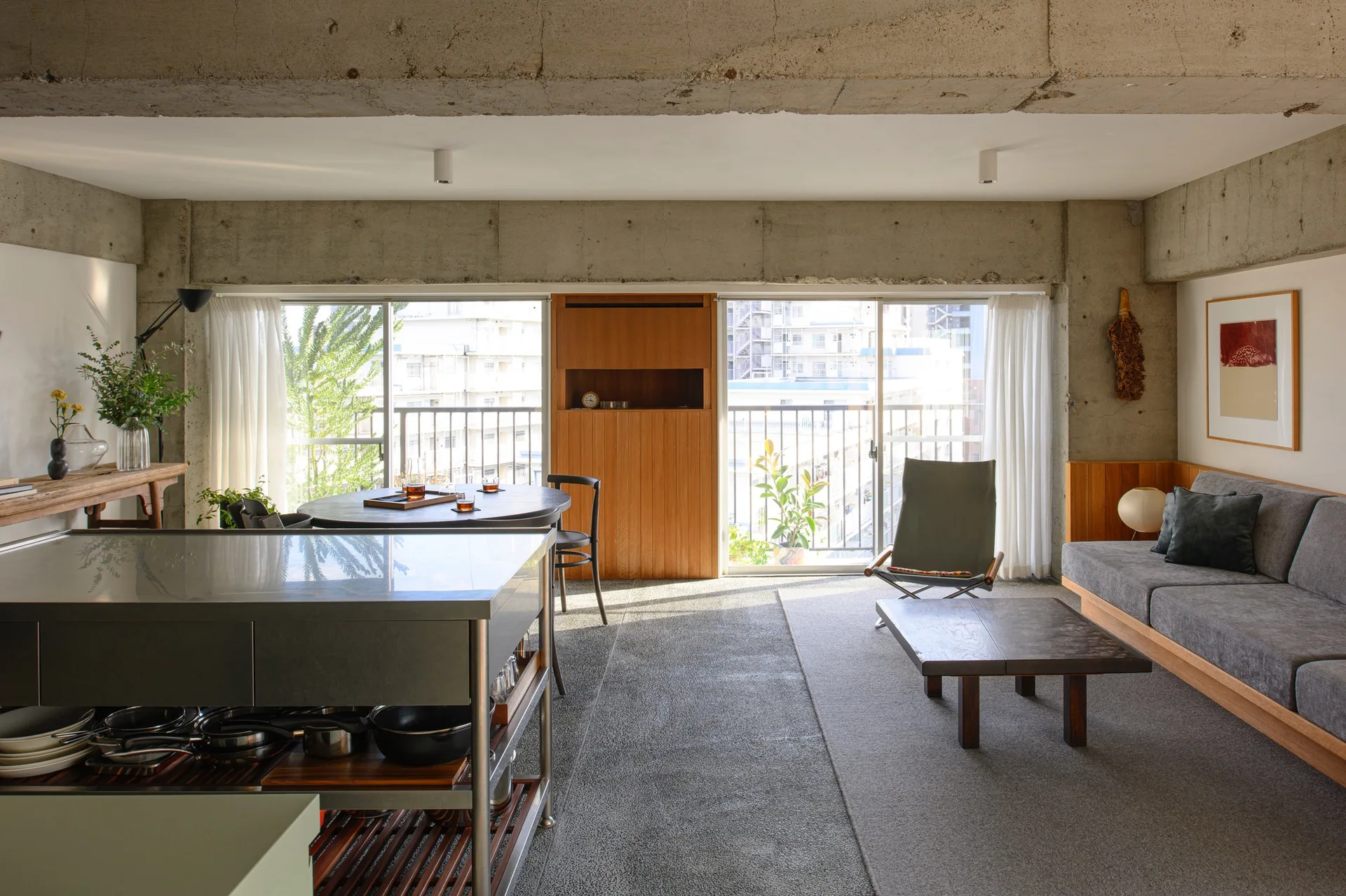 An open-concept living area features a grey sofa, a low wood coffee table, and an exposed concrete ceiling next to a stainless steel kitchen island.