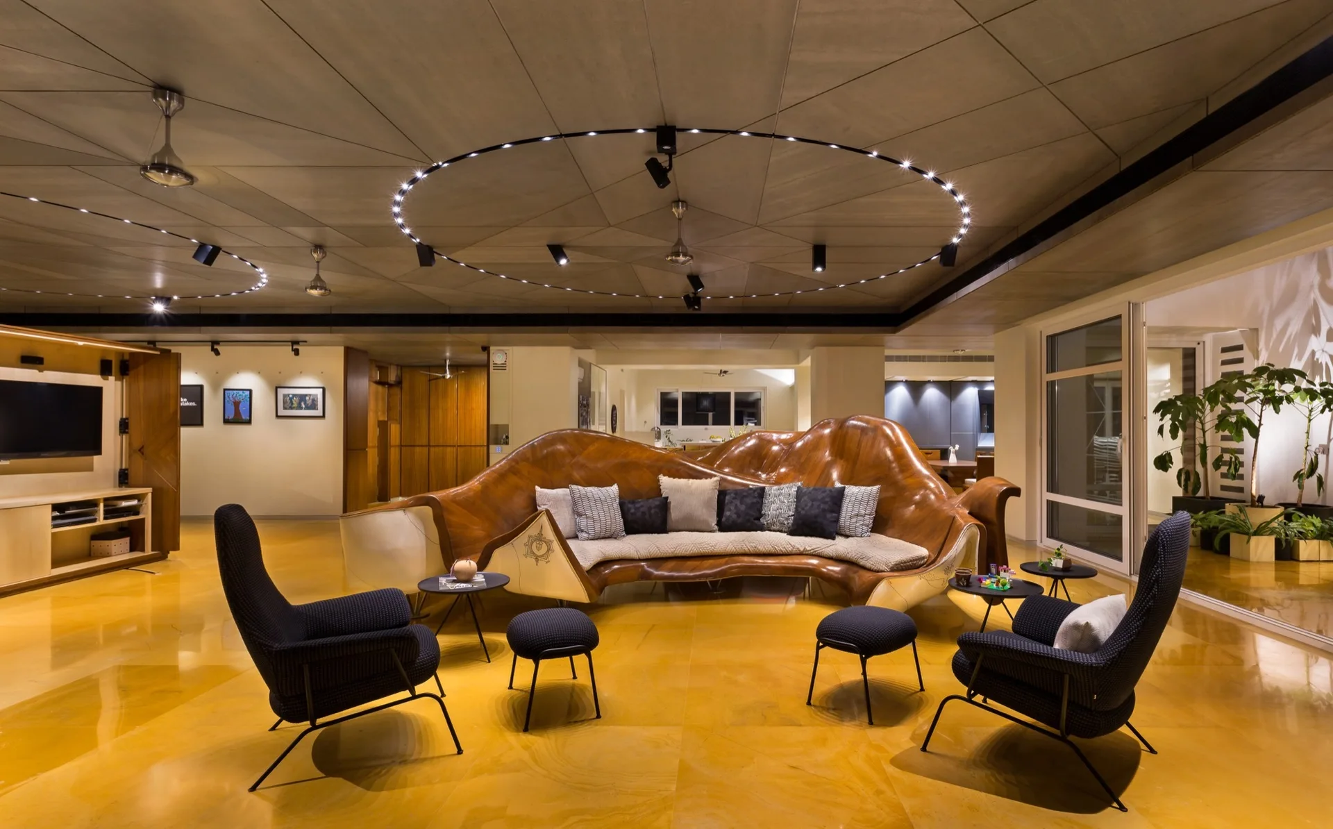 A spacious living room features a large curved wooden sculptural sofa, dark armchairs, yellow stone floors, and a circular ceiling light fixture.