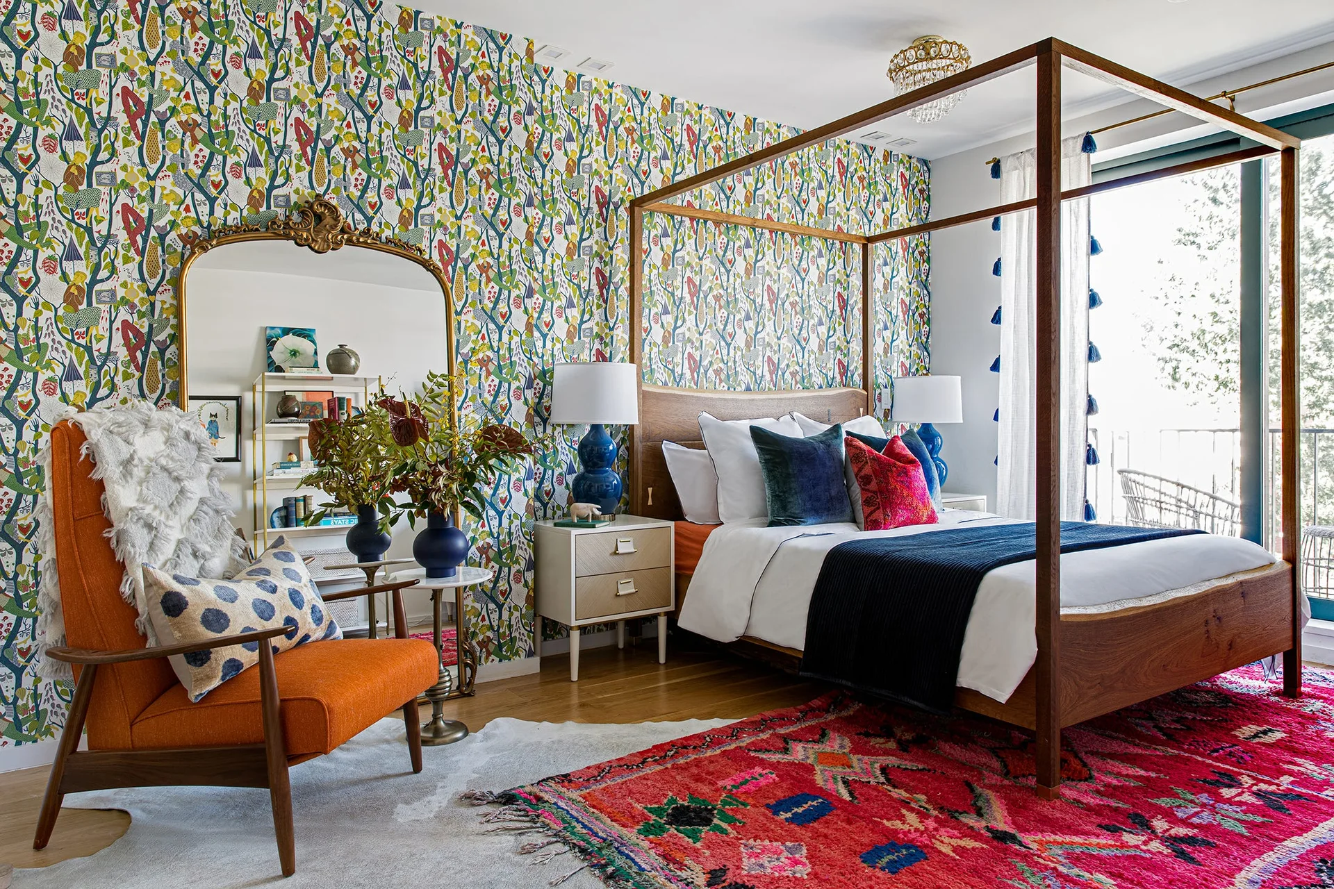 An eclectic bedroom features a wooden four-poster bed set against colorful botanical wallpaper and a red patterned rug.