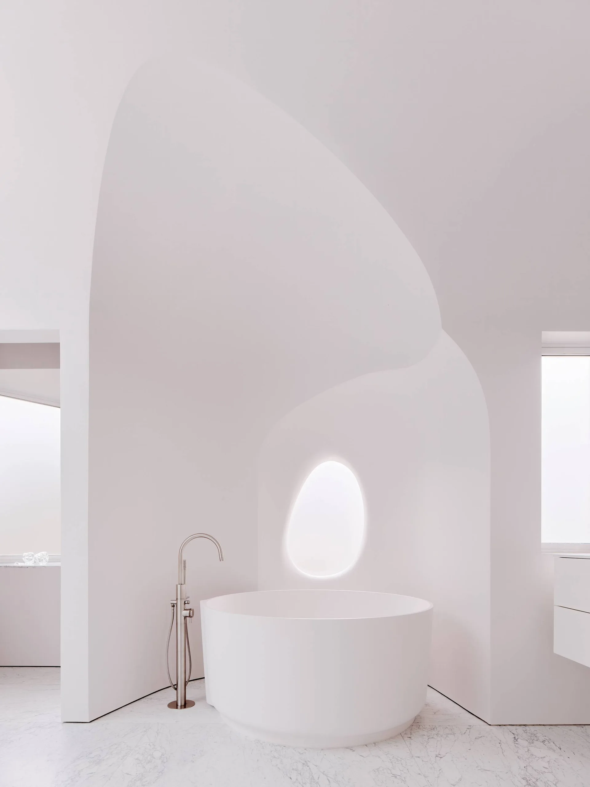A minimalist white bathroom with a circular soaking tub, floor-mounted faucet, curved architectural alcove, and veined marble flooring.