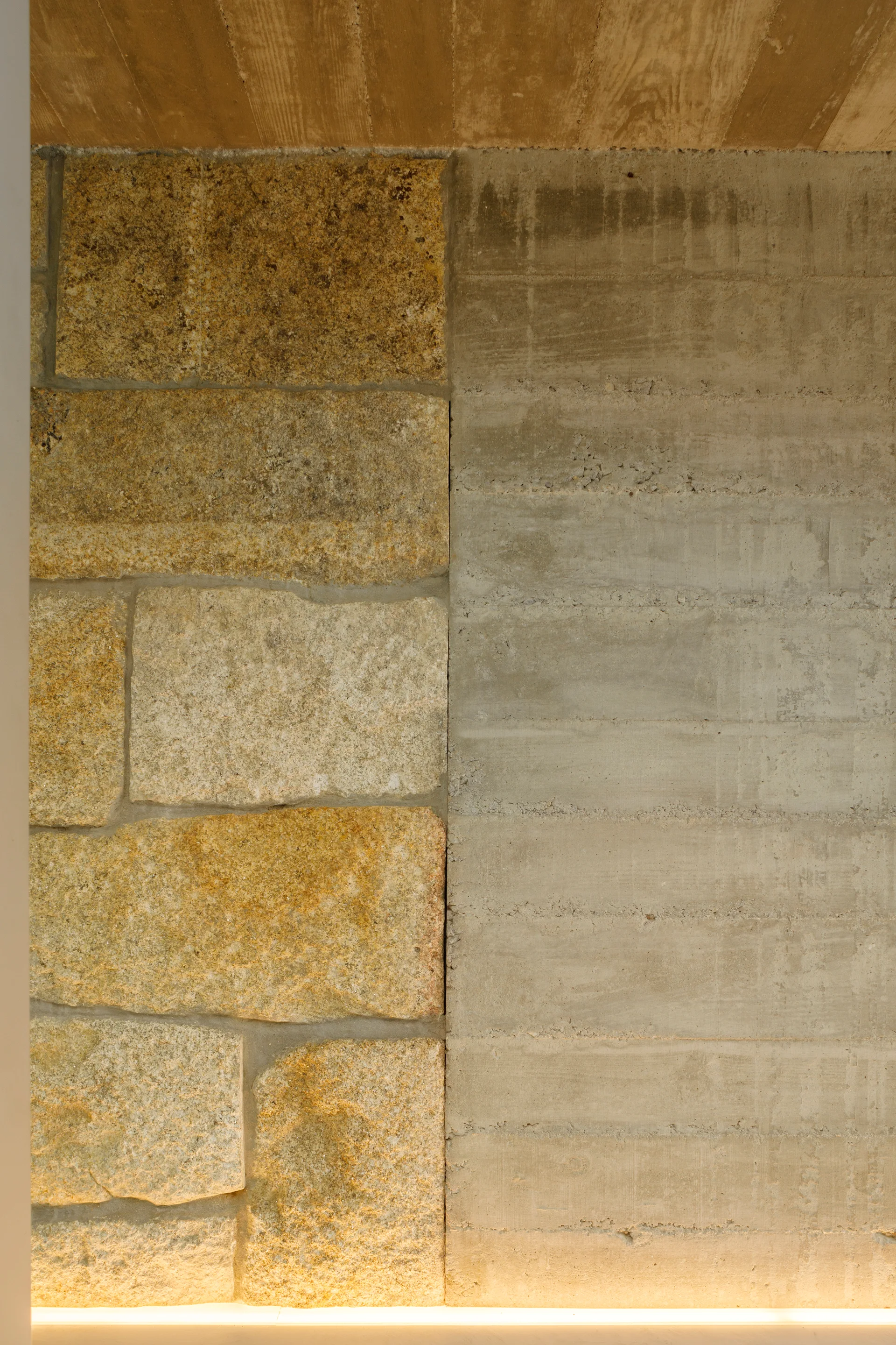 A wall featuring a vertical split between golden rough-cut stone and grey board-formed concrete, highlighted by a warm linear uplight at the base.