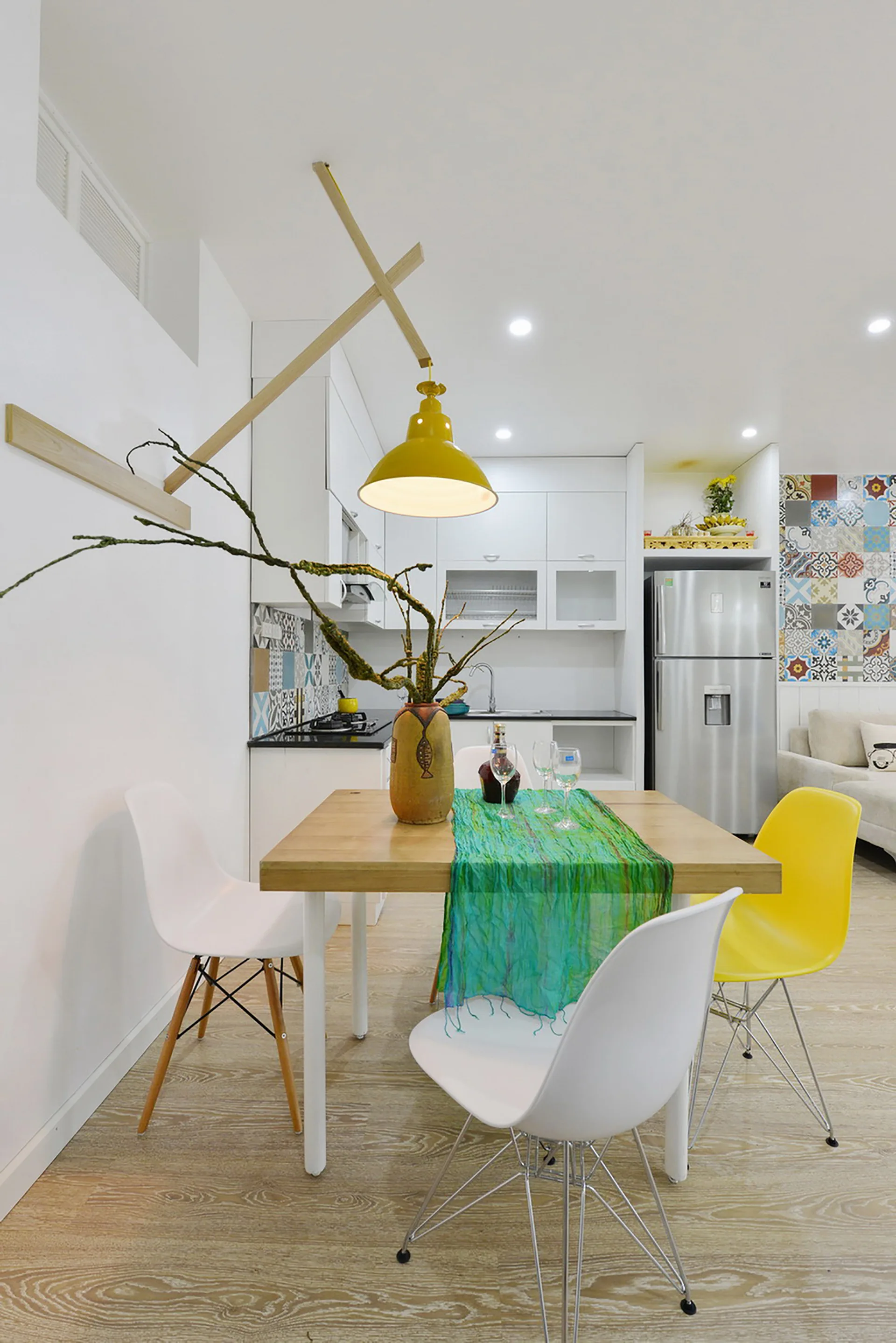 A wooden dining table with white and yellow chairs sits below a yellow pendant light in a white kitchen with a patterned tile backsplash.