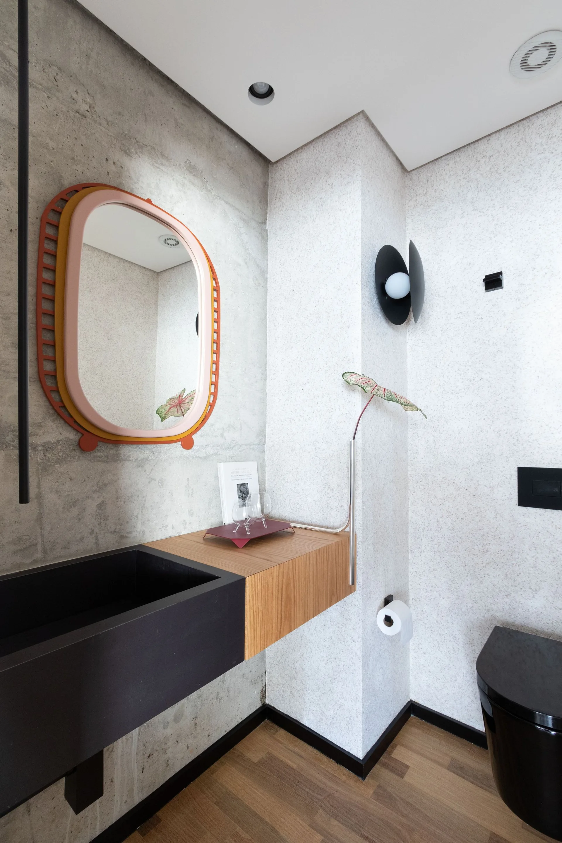 A modern powder room features a concrete wall, a floating wood vanity with a black sink, and a colorful rounded mirror.