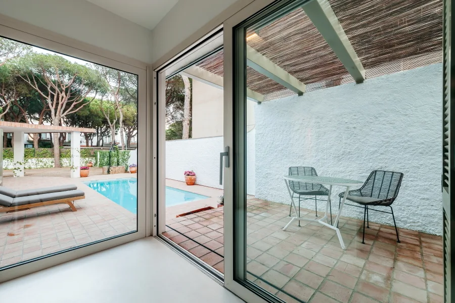 A view through glass doors looking out at a terracotta patio with a bistro set under a bamboo pergola and a swimming pool in the background.
