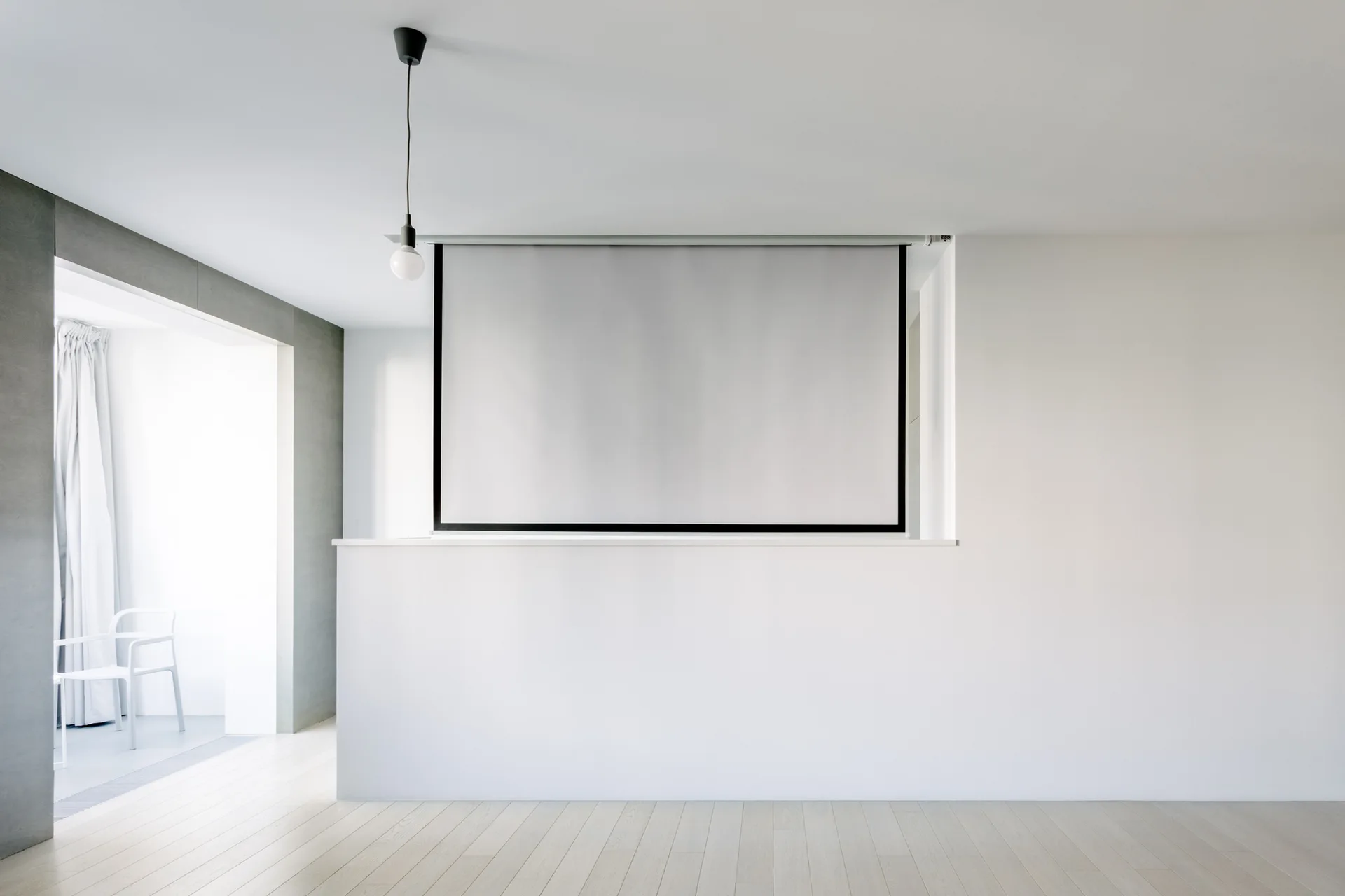 A minimalist room features a retractable projector screen mounted above a white half-wall partition, pale wood floors, and a black pendant light.